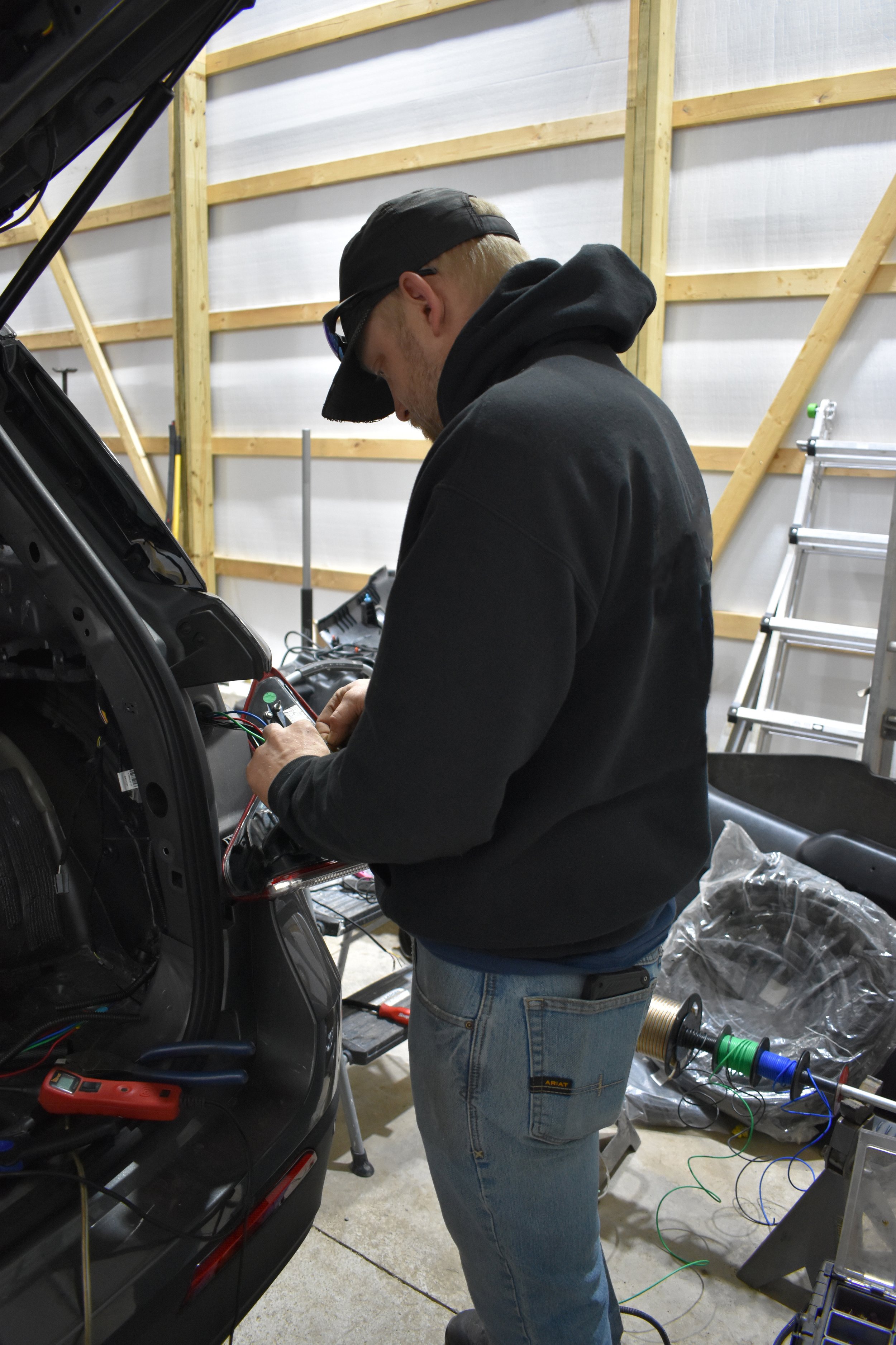 Man working on wiring inside an electric vehicle in a workshop with a ladder and tools around.