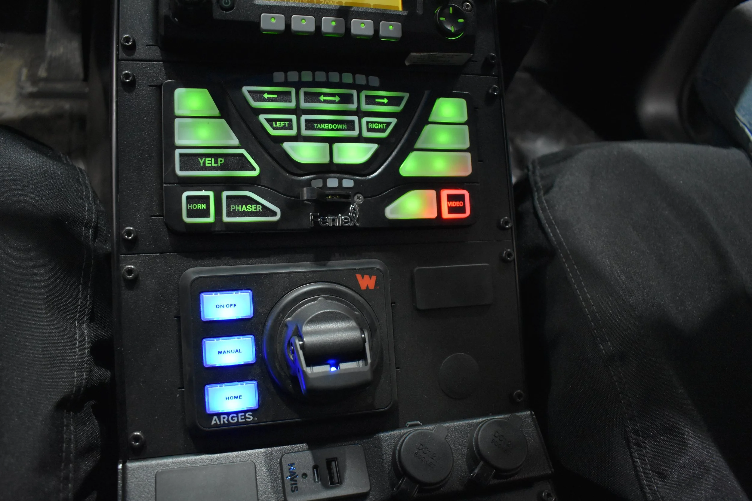 Close-up view of a control panel with illuminated buttons, including directional arrows, labeled left, right, takedown, Yelp, horn, phaser, video, and home, with some additional buttons and switches.