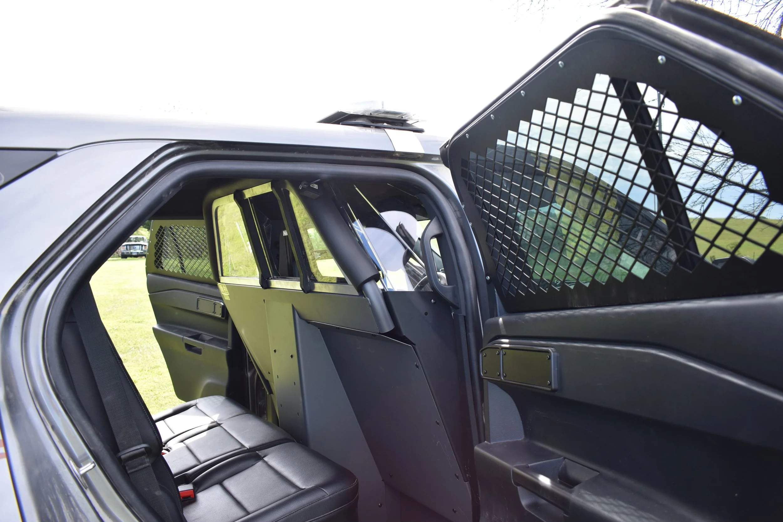 Close-up of a police or security vehicle's interior, showing the backseat with black leather seats, a partition with a protective grid, and a view through the open door revealing a grassy area outside.
