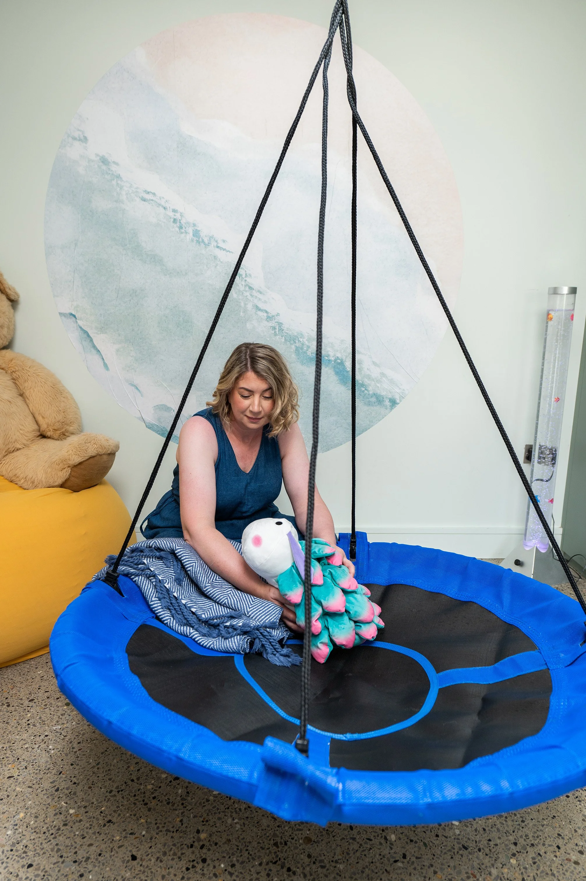 A woman sitting beside a blue and black round swing, holding a stuffed toy.