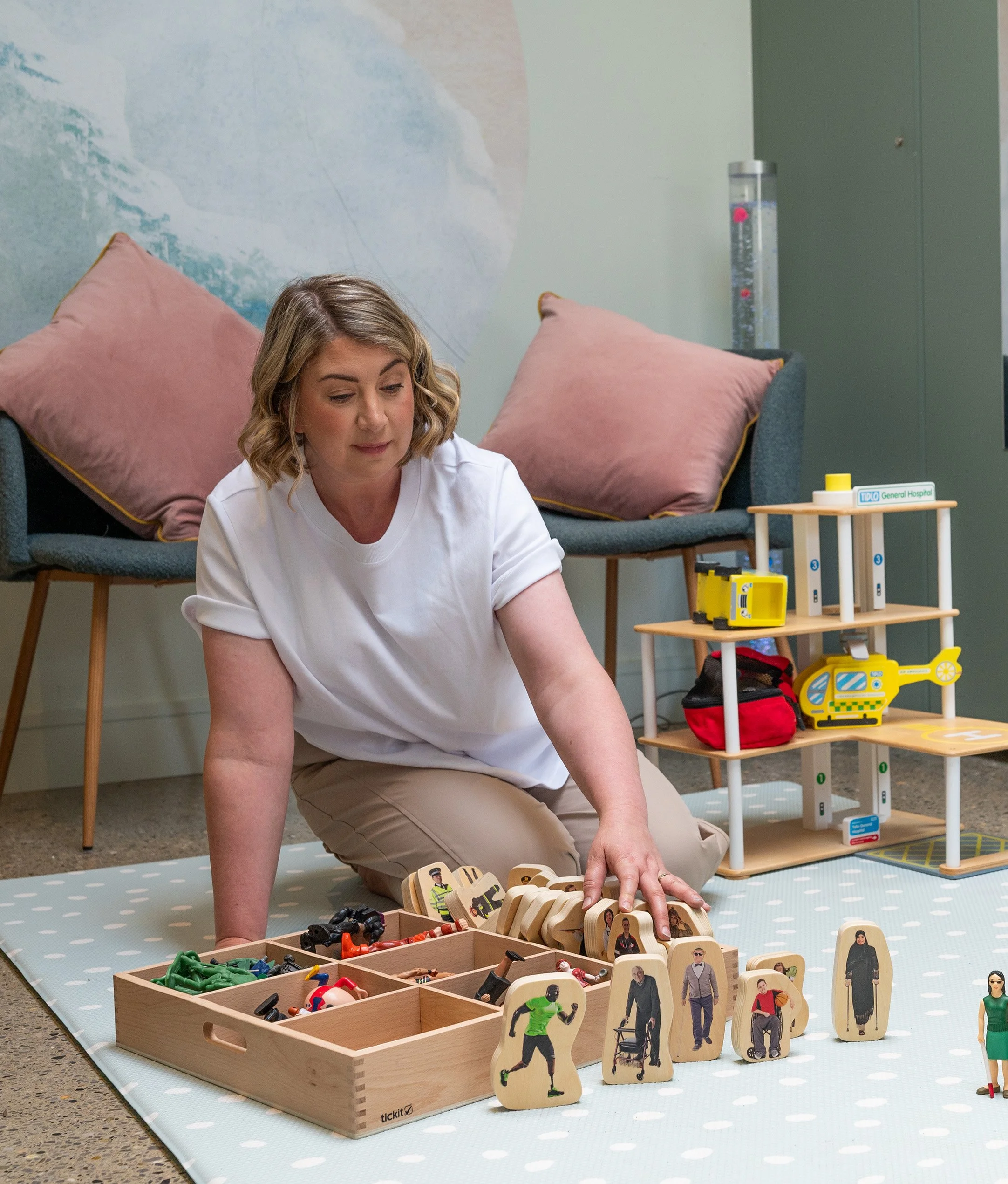 A woman in a white shirt plays with wooden toys seated on a light blue mat in a room with pink pillows on the chairs.
