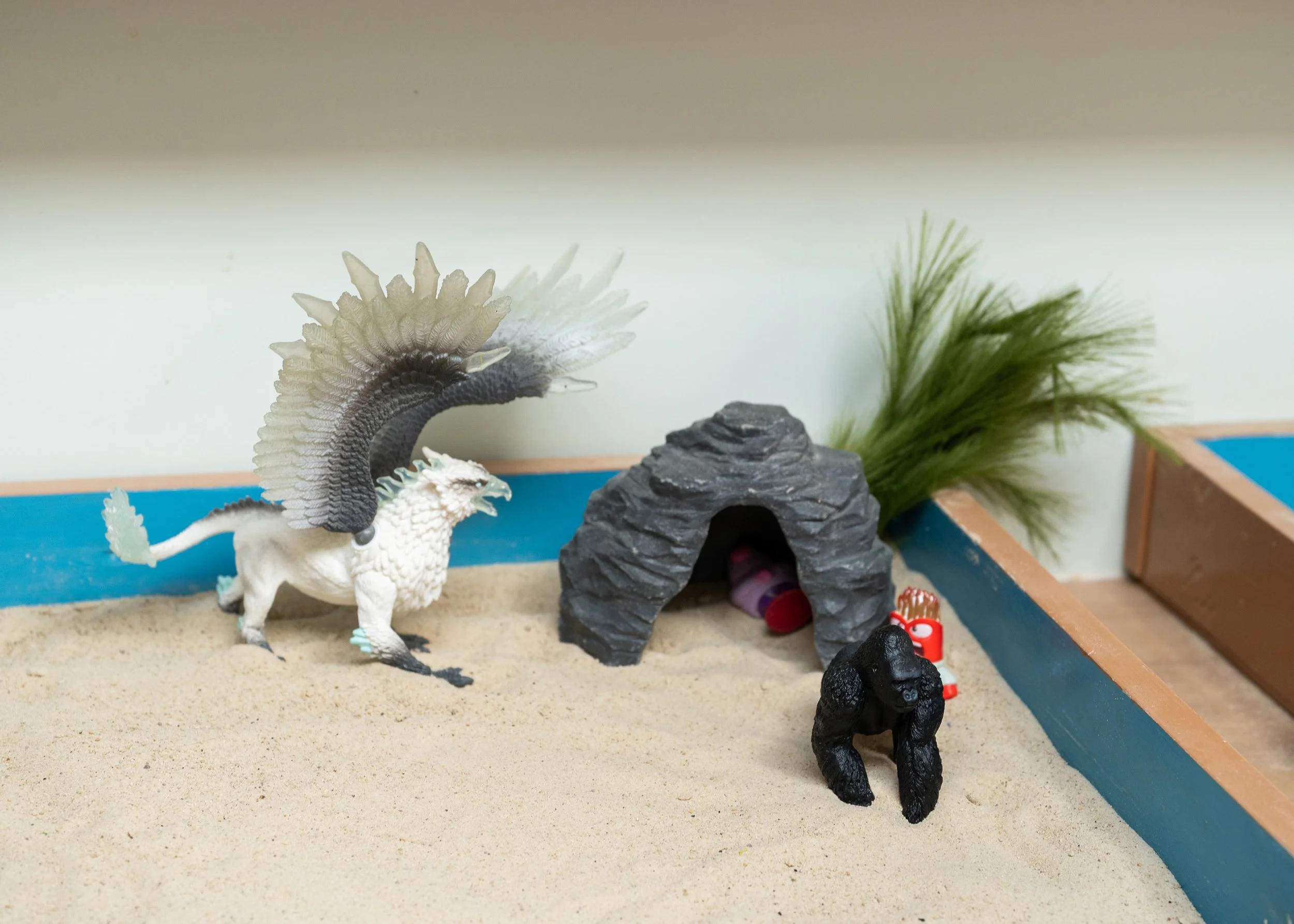 A toy setup featuring a white dragon figurine, a rock cave, and some artificial plants on a sand in a framed square.