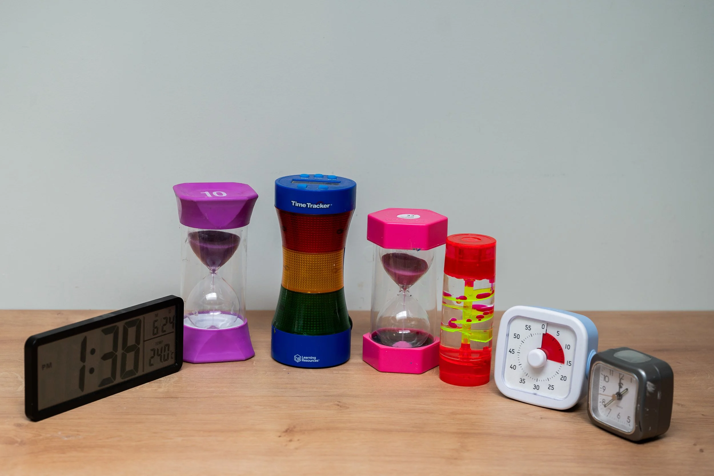 Various timers and clocks on a wooden table, including digital and analog types, with three hourglasses, against a white wall.