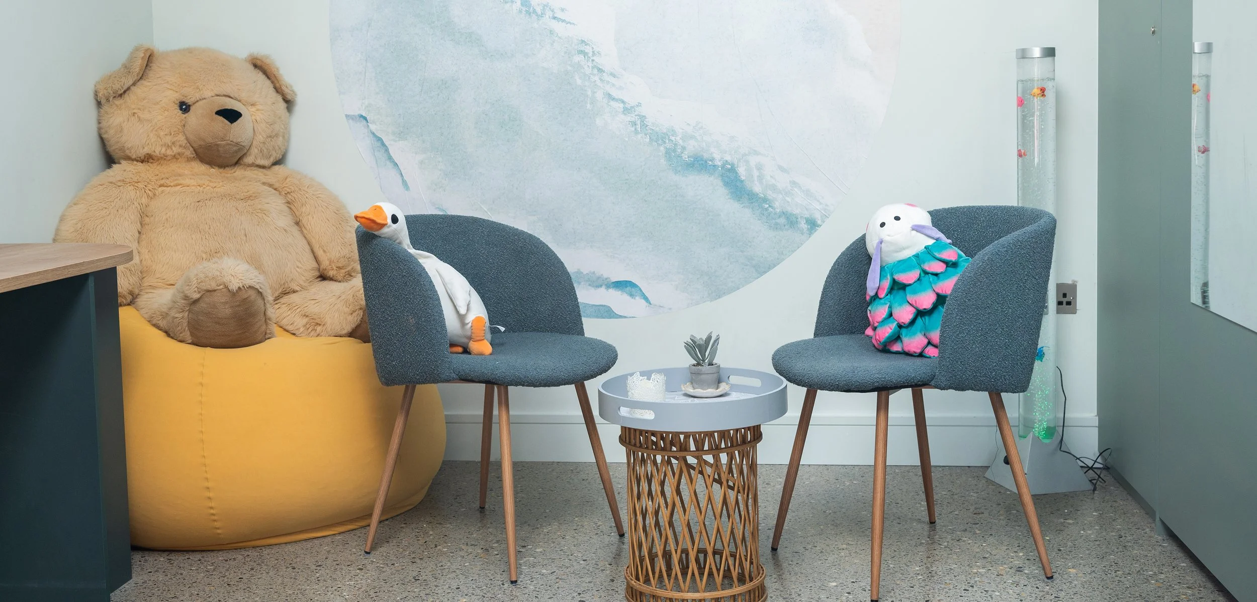 A seating area with two gray chairs, a small round table with a plant, a large yellow bean bag with a plush teddy bear.