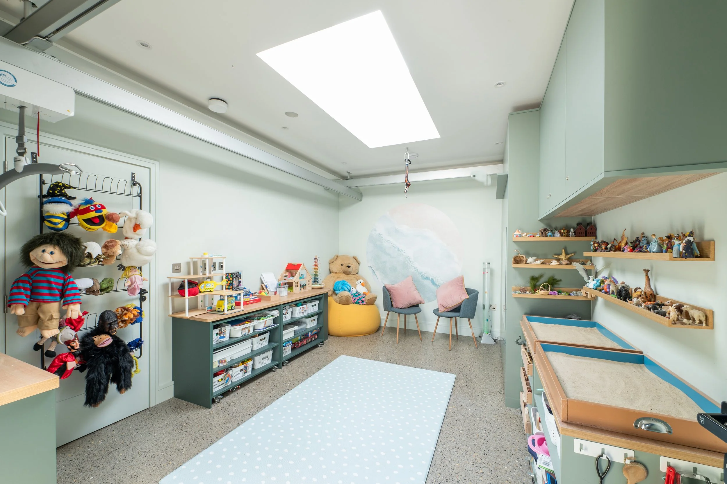 A room with shelves of puppets, stuffed animals, and toys, two chairs with pink pillows, a large yellow beanbag, and a light green wall with a mural of a wave.