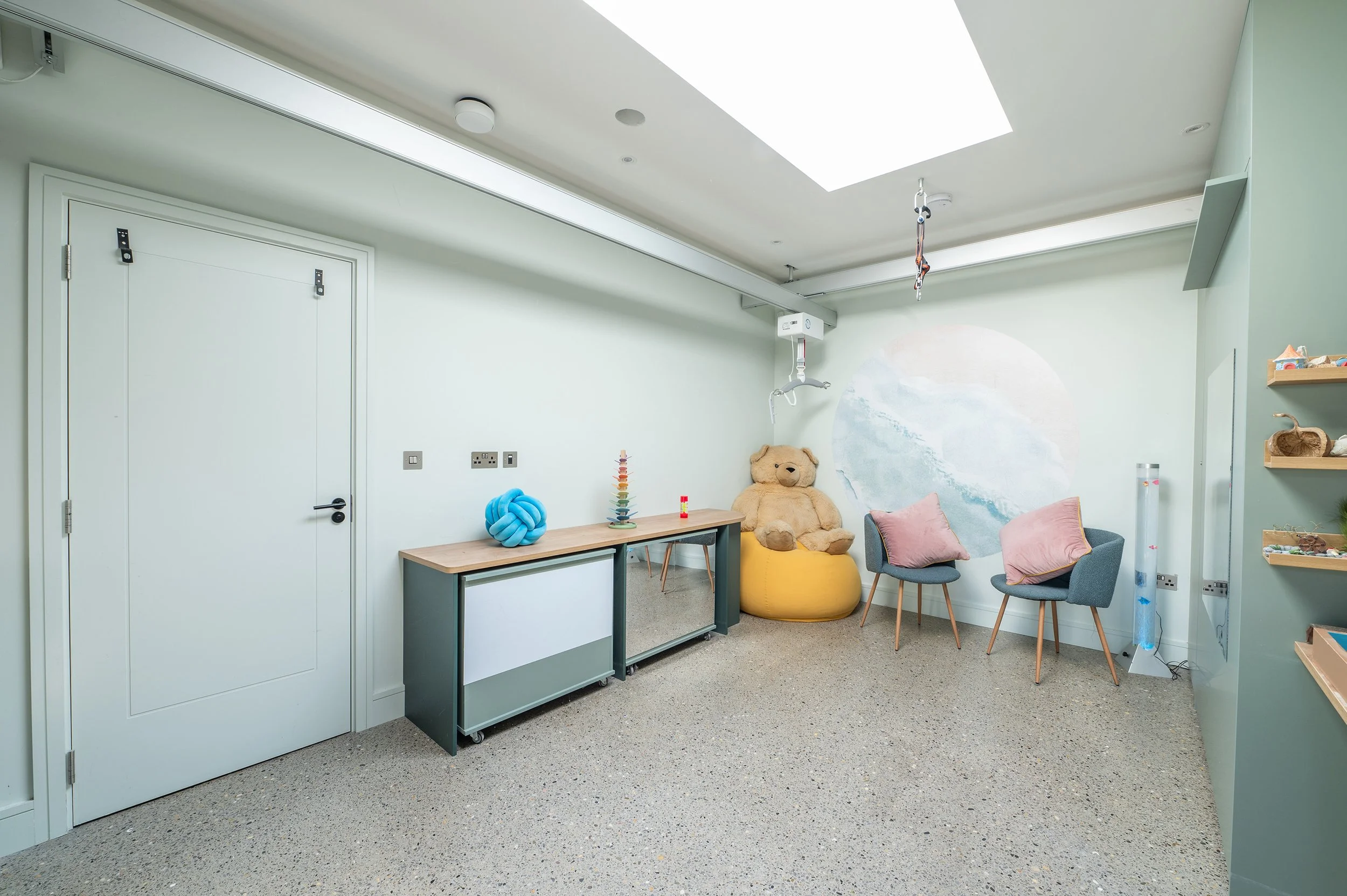 A room with a mobile mirror cabinet, giant teddy bear, yellow bean bag, and two grey armchairs against a soft mountain mural.