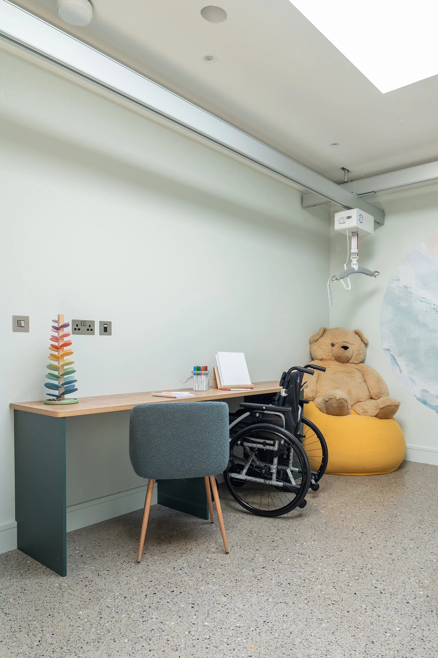 A room with a desk, a chair, a wheelchair, a large teddy bear sitting on a yellow beanbag.