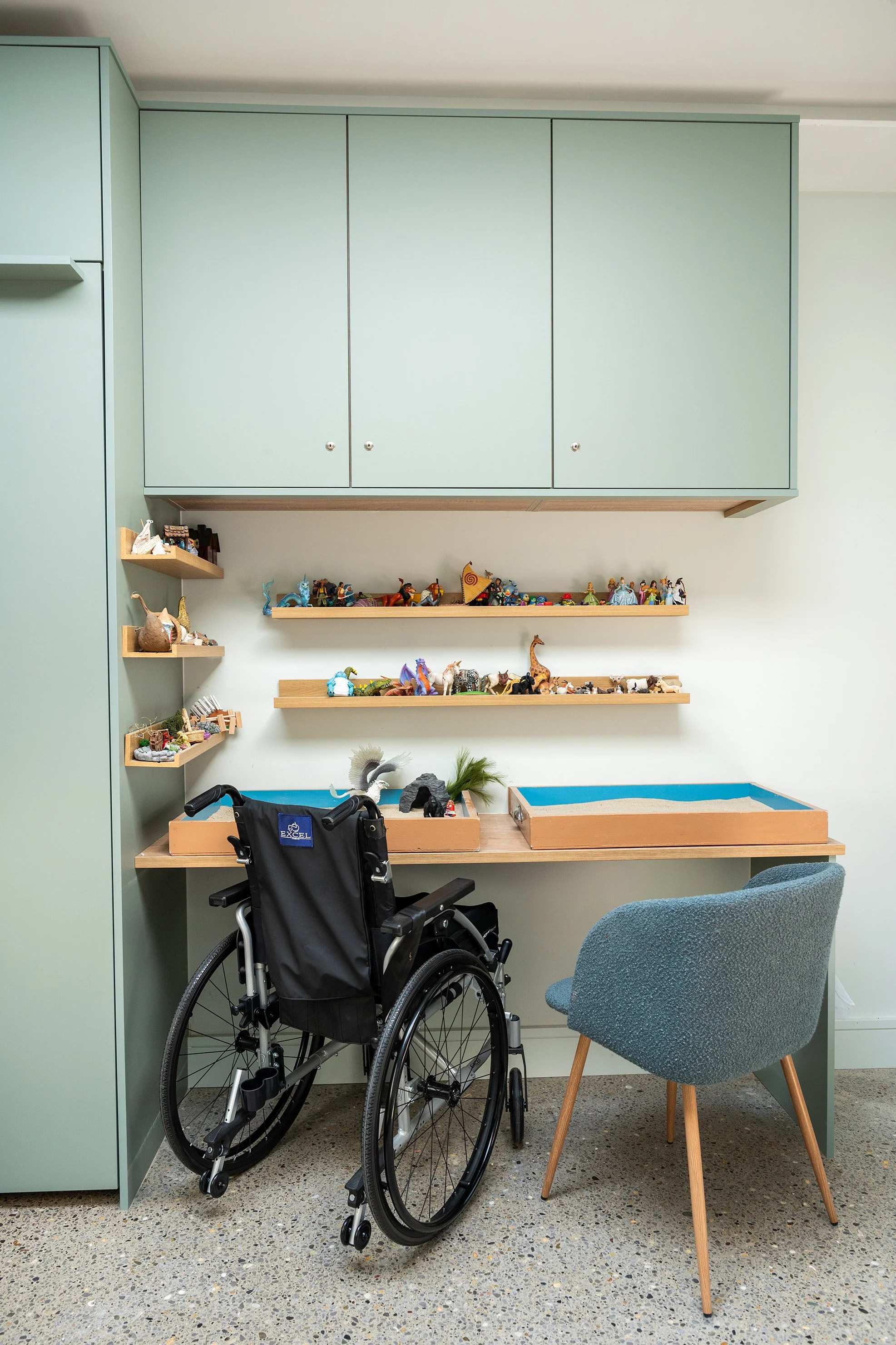 Room with light green cabinets, wooden shelves filled with small figurines and toys, a wheelchair, a grey chair, and a table.