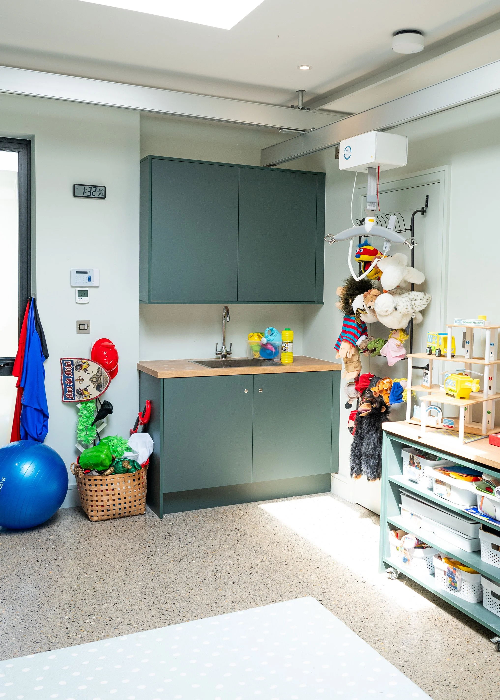 A room with a green kitchenette unit, ceiling hoist with hanging plush toys, exercise ball, and shelving stocked with children's toys and storage bins.