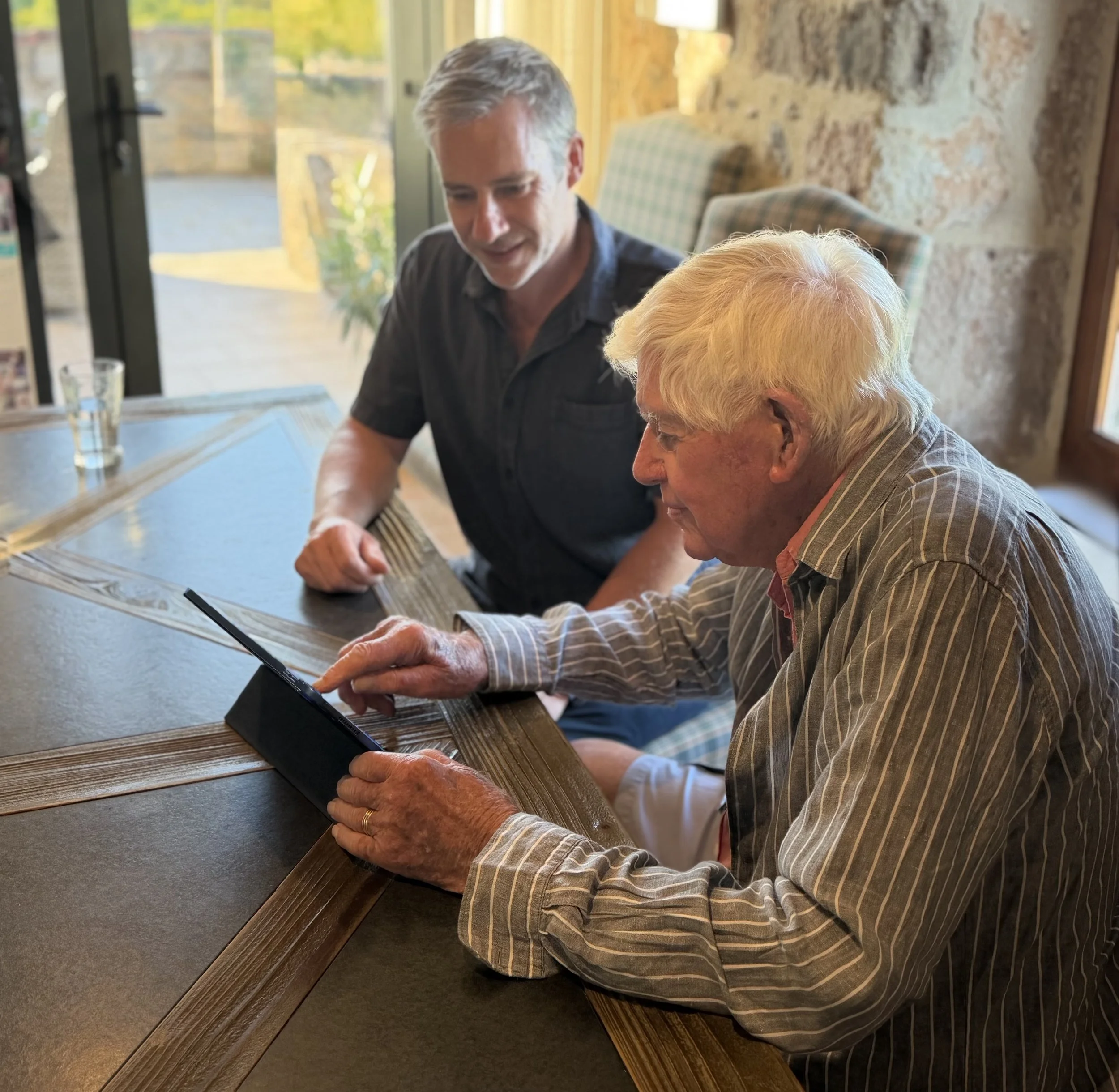Two men are sitting at a wooden table inside a restaurant or cafe, engaged in conversation. The background shows large windows with a view of greenery outside.