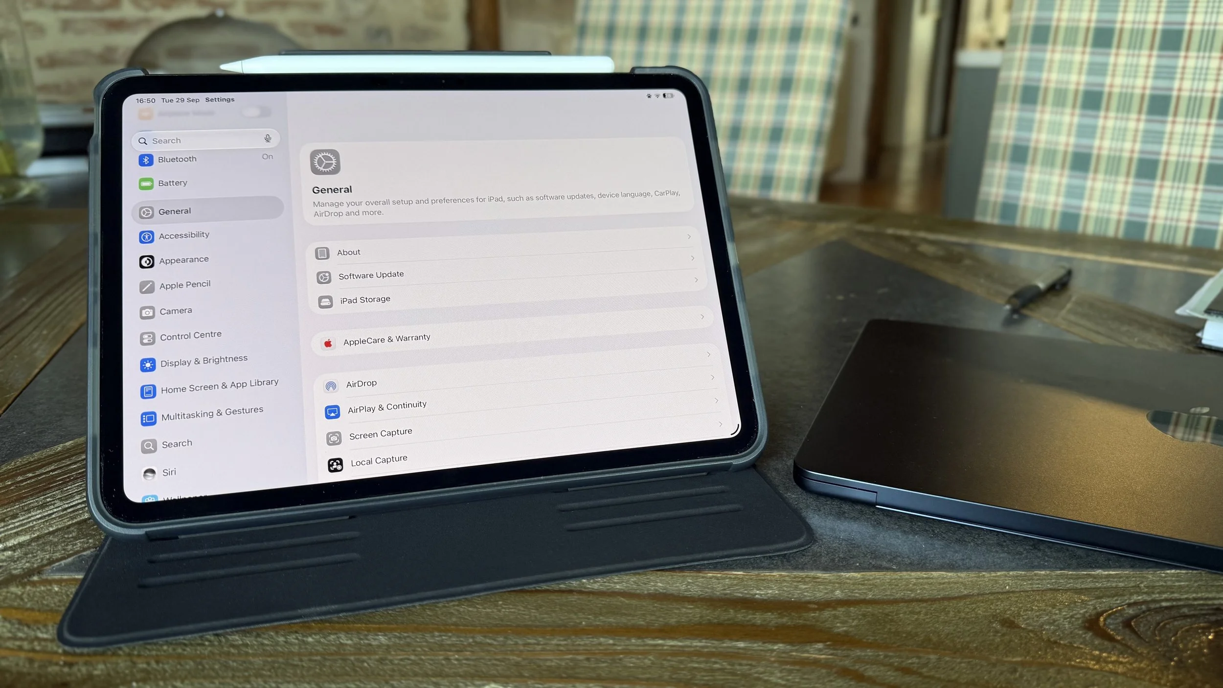 An iPad in landscape mode displaying settings, on a wooden table with a pen and a laptop nearby.