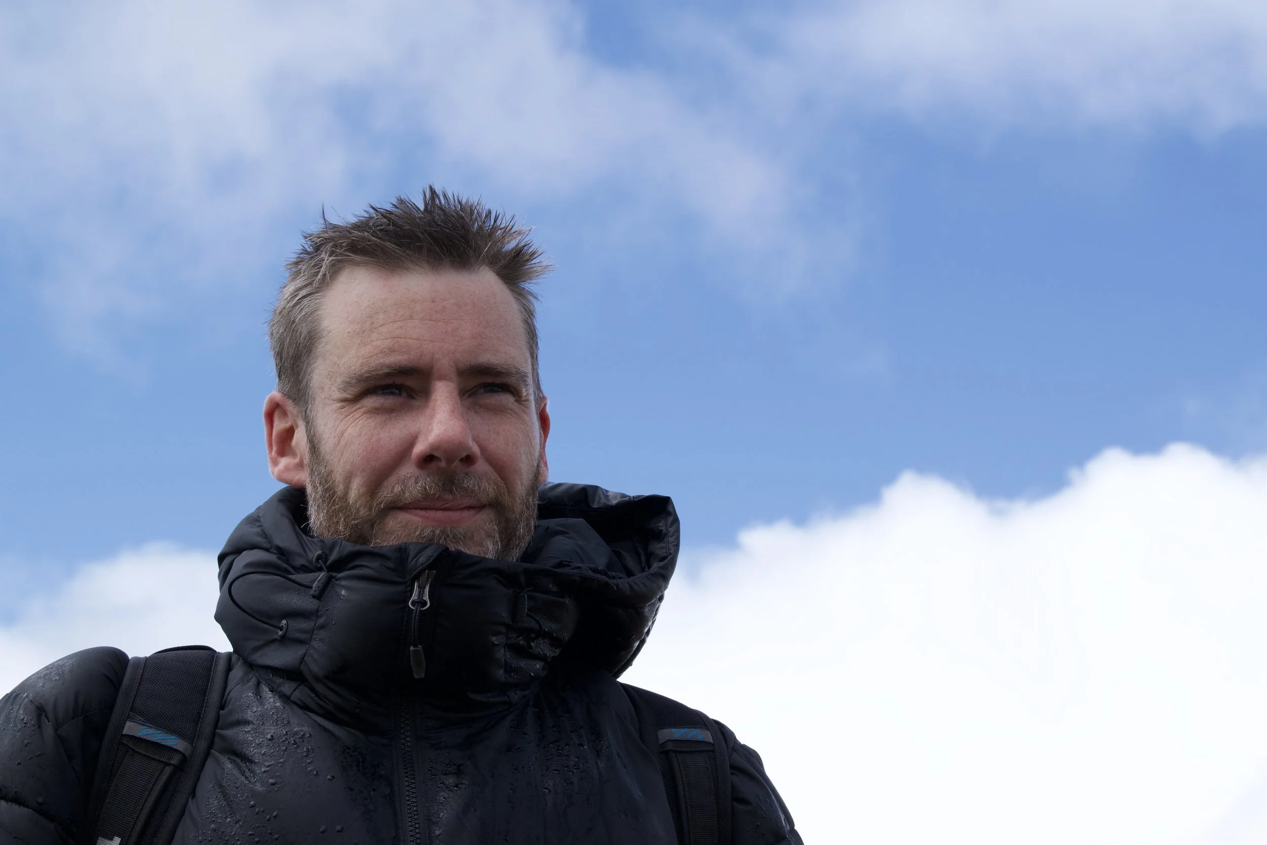 A man with brown hair and a beard wearing a black jacket and backpack outdoors against a blue sky with clouds.
