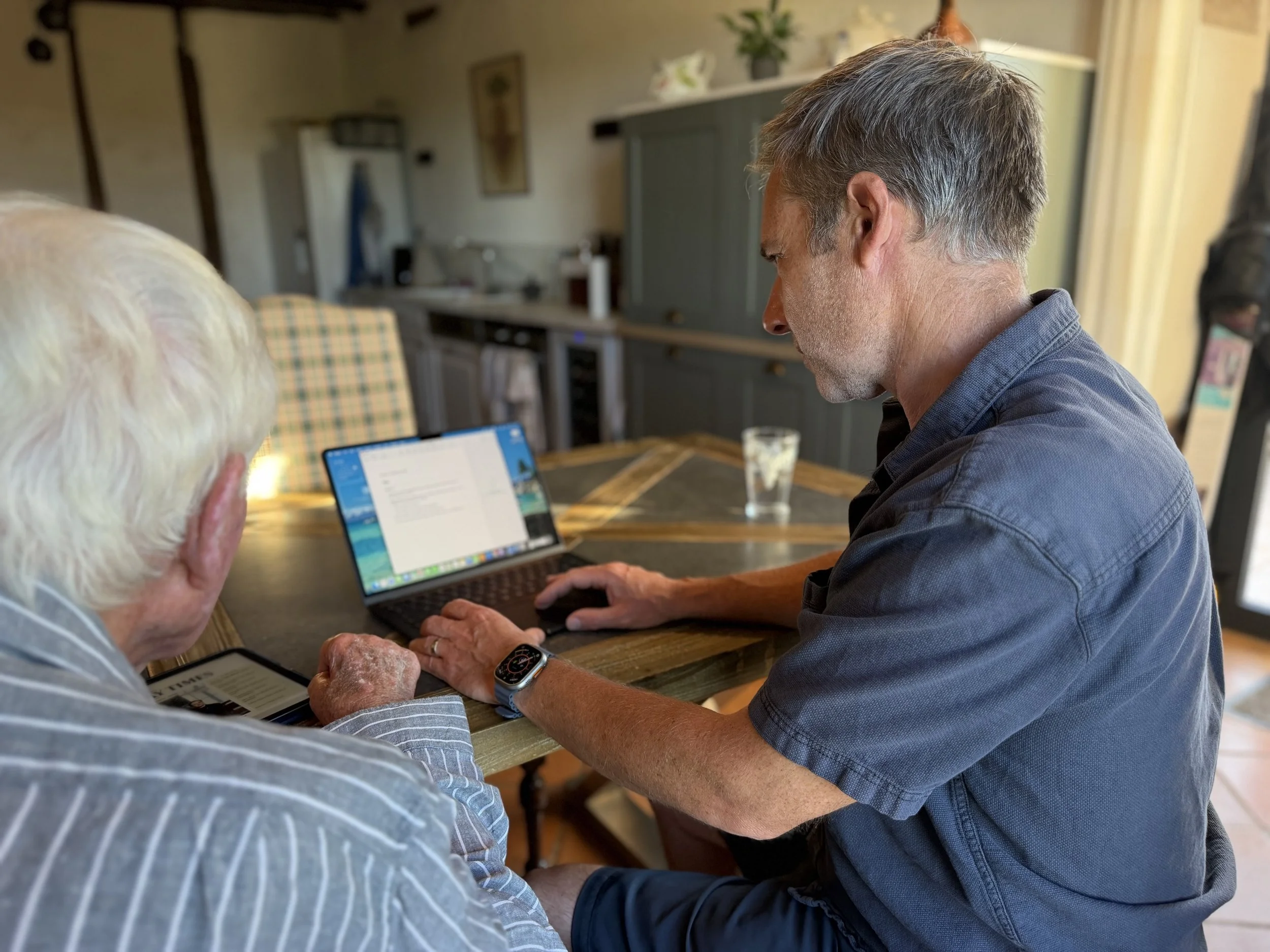 Two men sitting at a wooden table, using smartphones and a laptop in a cozy home setting. One man wears an Apple Watch