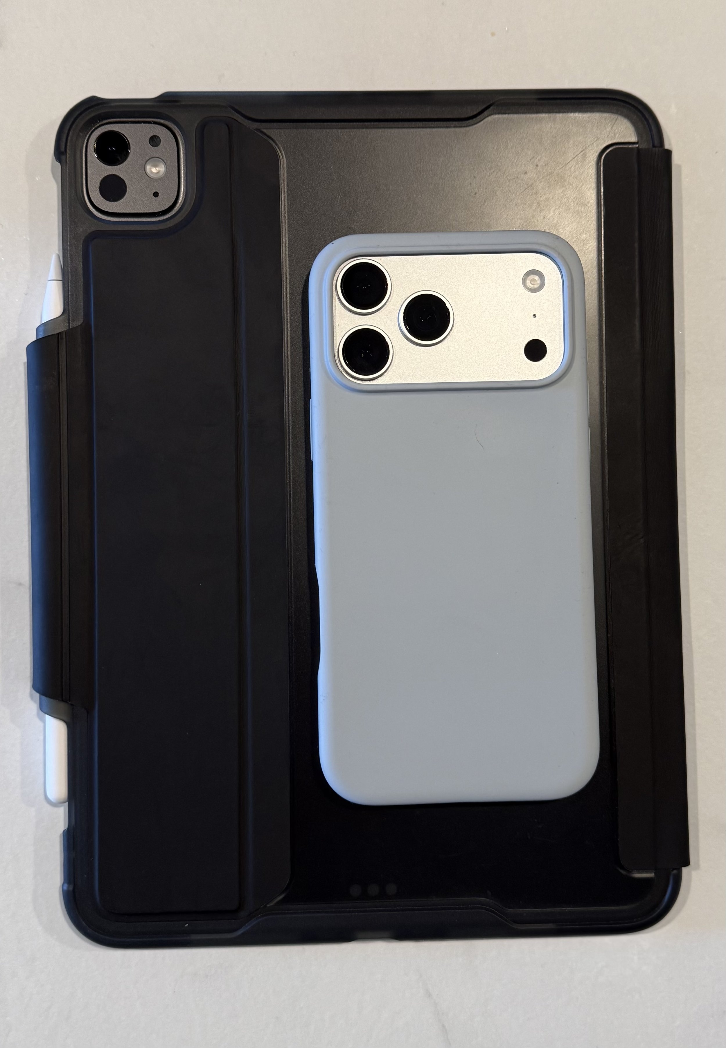 A smartphone with triple camera lenses and flash on the back, placed on top of a black iPad case with Apple Pencil Pro. The case has a compartment with a closed flap.