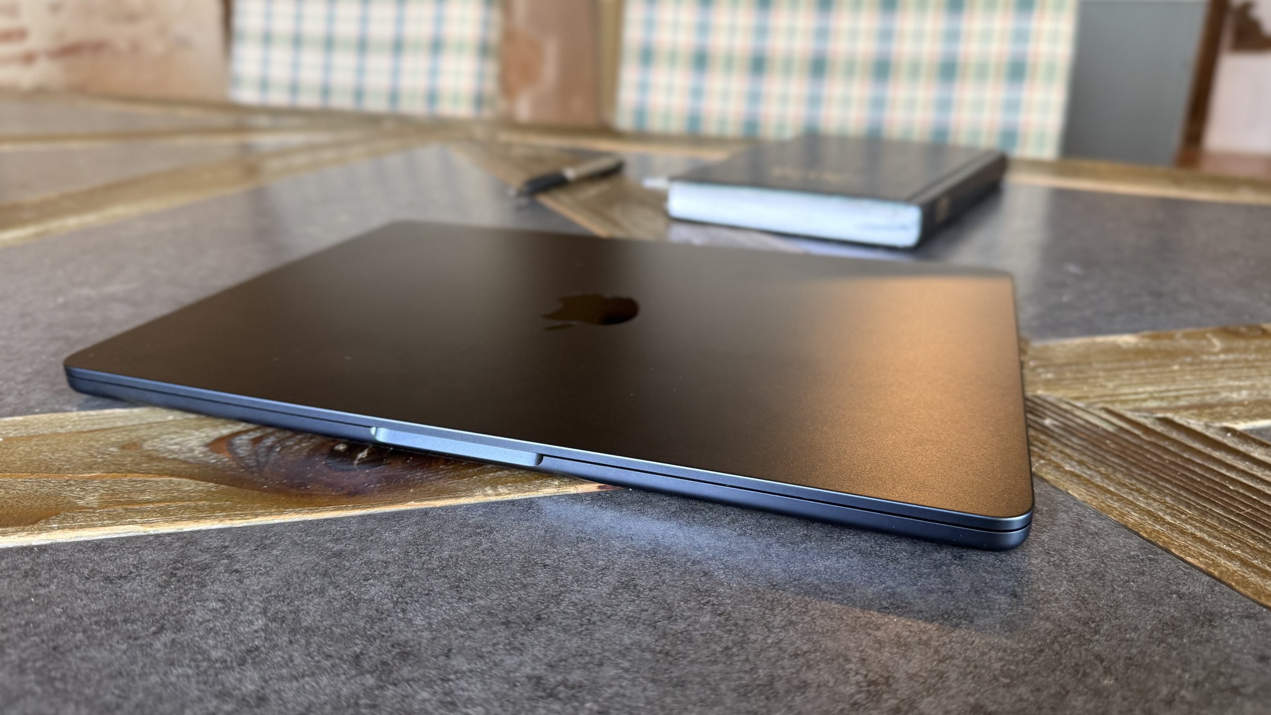A closed black laptop placed on a wooden table with a small notepad and a pen in the background.
