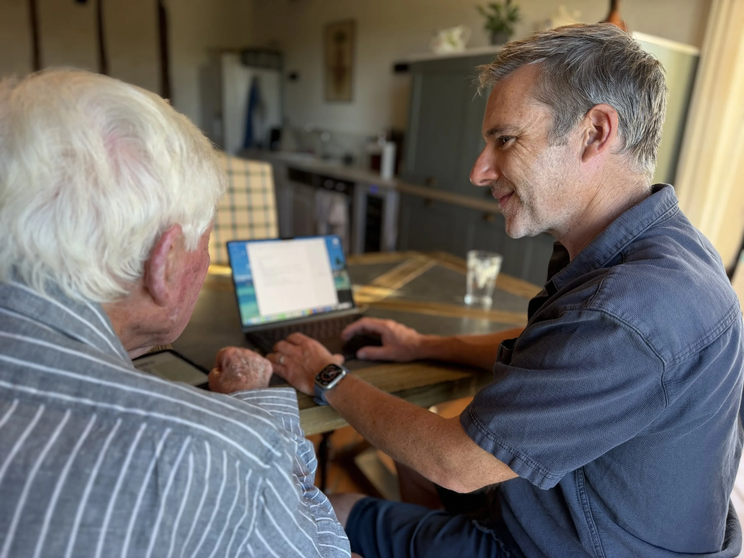 A man with gray hair smiling and looking at an elderly person with white hair, sitting at a wooden table with a laptop and a glass of water, in a cozy room with plants and decorations.