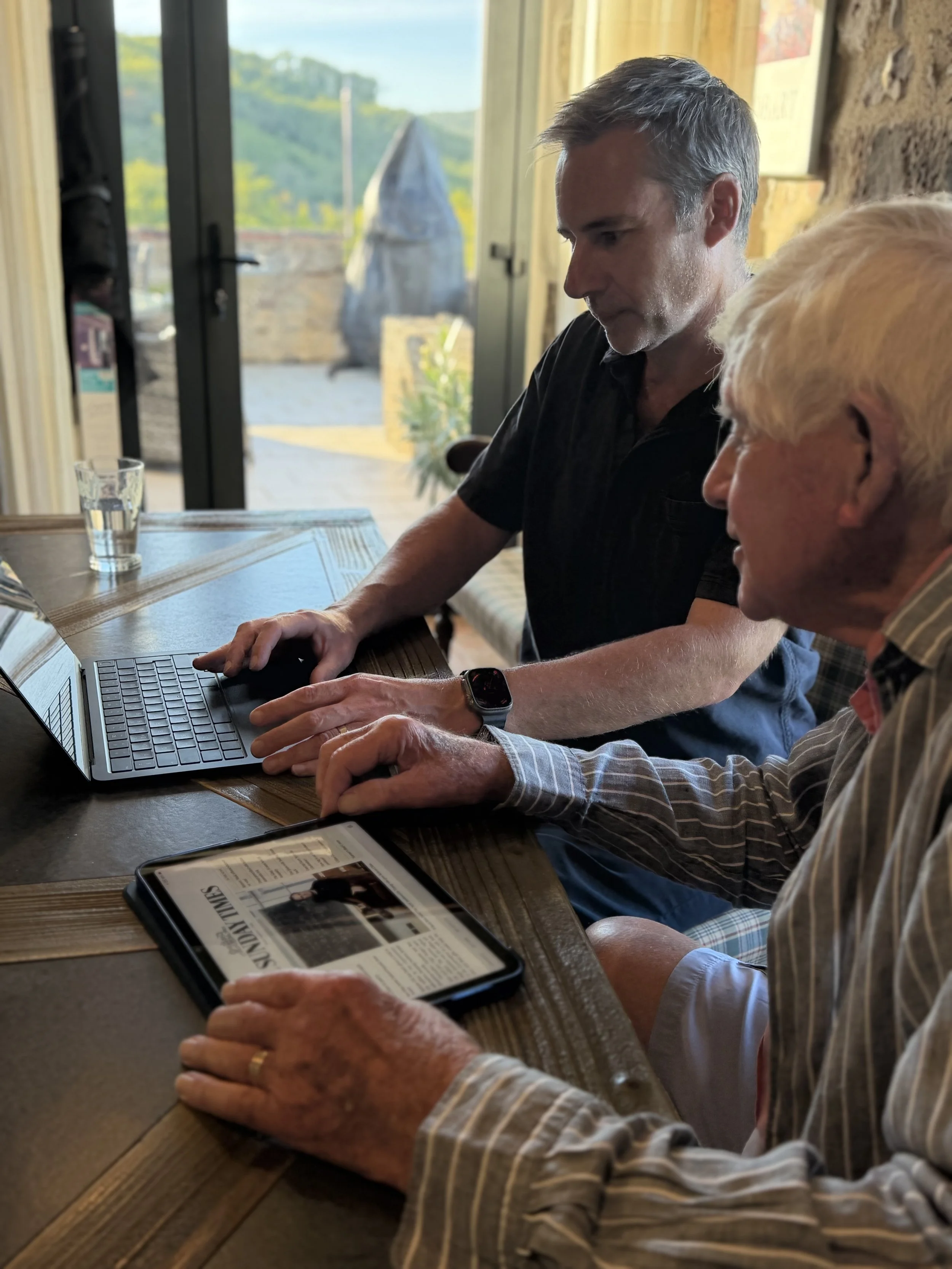 Two men sitting at a wooden table indoors, looking at a laptop screen. The man in the foreground is older, the man in the background looks friendly. An iPad looking at a newspaper is on the table.