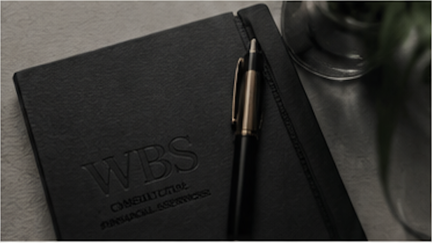 A black WBS construction notebook with a black and gold pen resting on top, placed on a gray surface near a plant.
