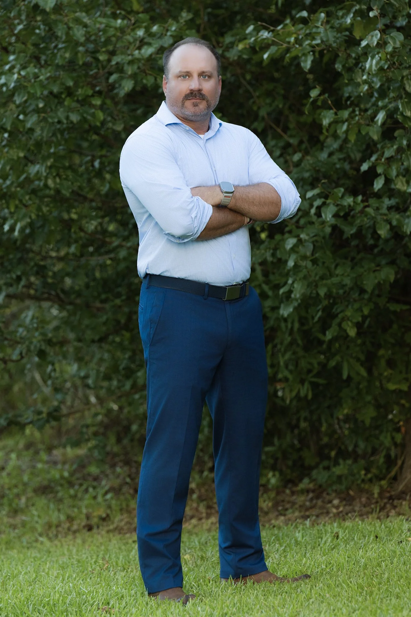 A man standing outdoors with arms crossed in front of green leafy background, wearing a light blue button-up shirt, dark blue trousers, and a smartwatch.