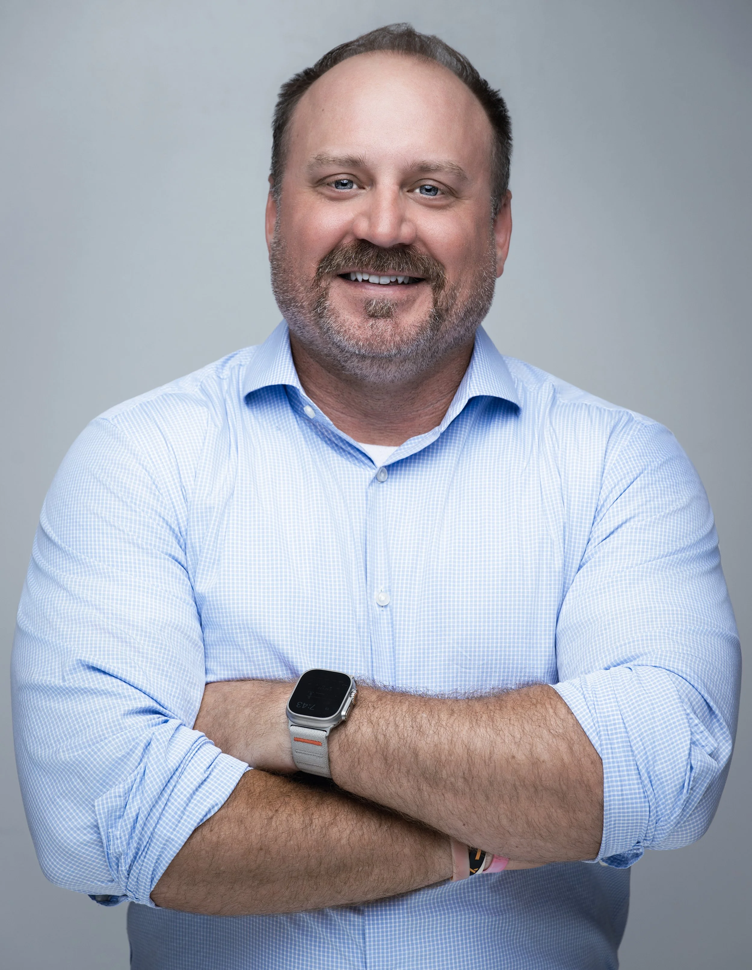 Portrait of a smiling middle-aged man with a beard and mustache, wearing a light blue dress shirt and a smartwatch on his left wrist, standing against a gray background.