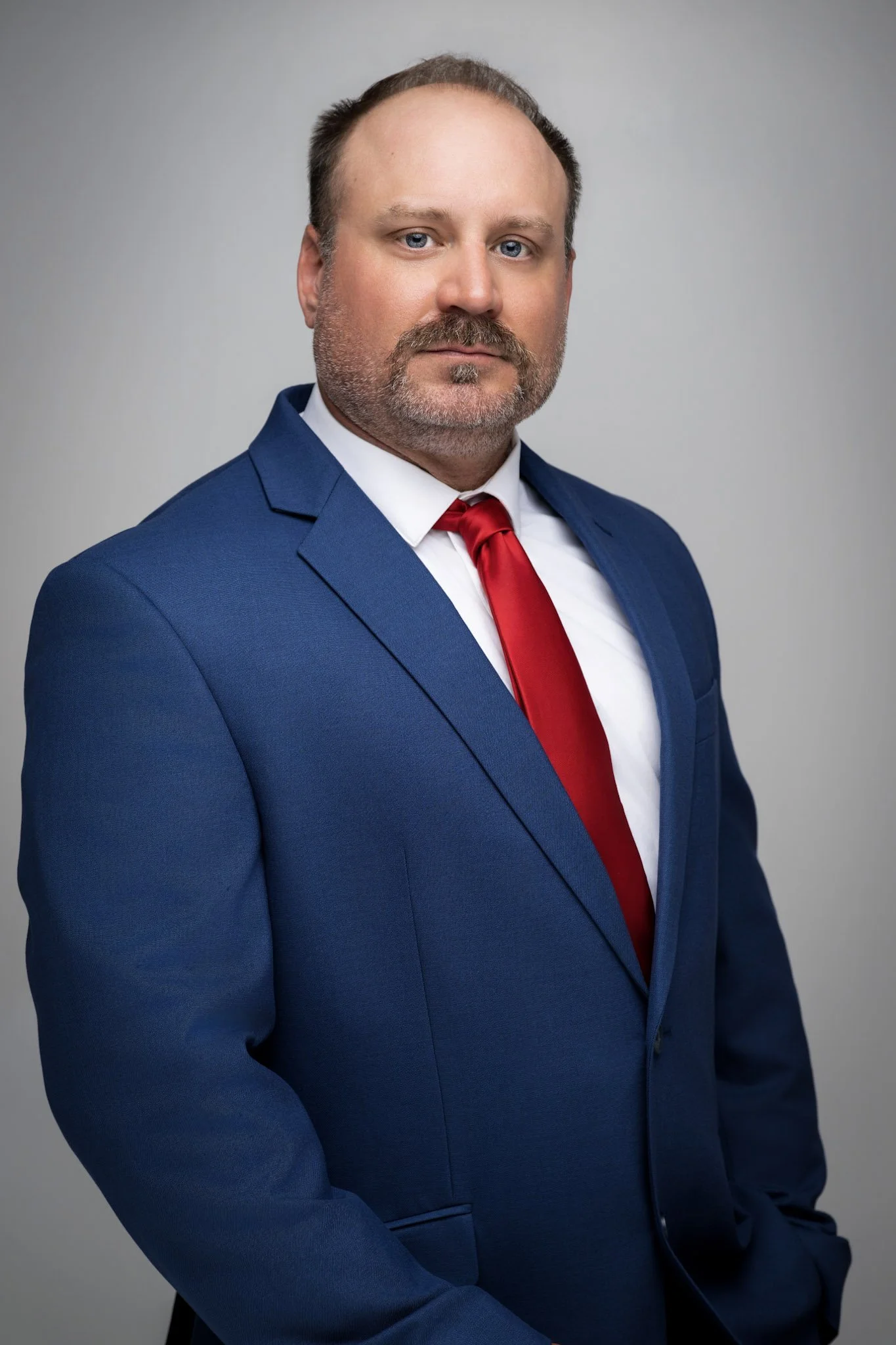 Portrait of a man with light skin, blue eyes, brown hair, and a beard, wearing a blue suit, white shirt, and red tie, against a plain gray background.