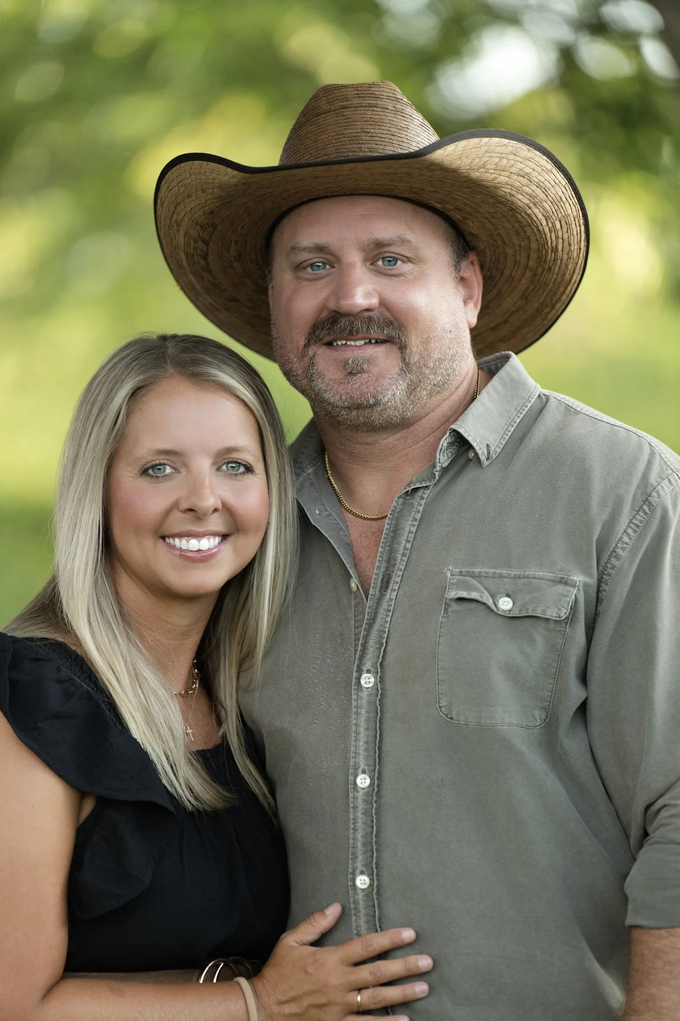 A man wearing a large straw cowboy hat stands next to a woman with long blonde hair, both smiling, with a green, blurred outdoor background.