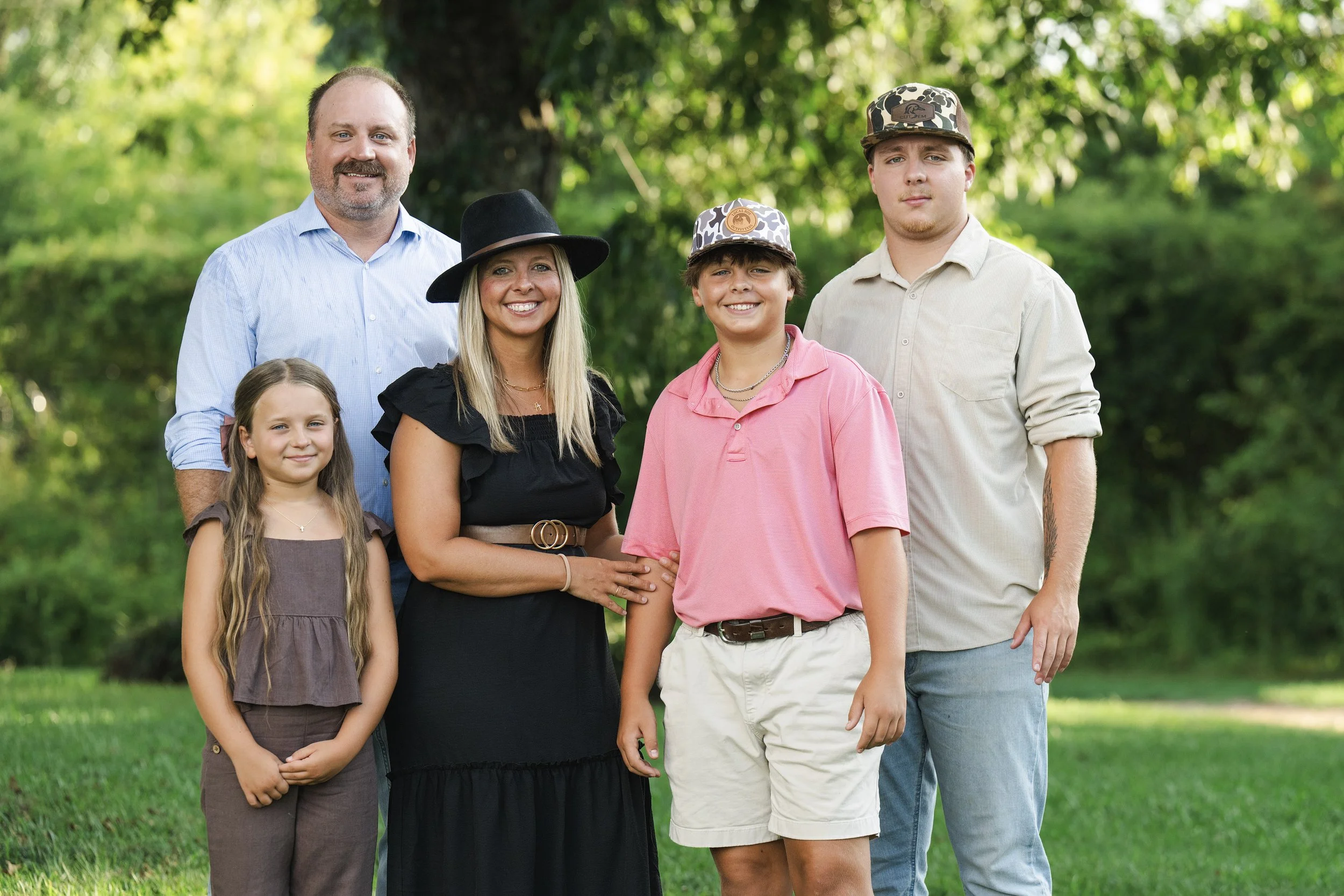 A diverse group of six people standing outdoors in a park with green trees in the background. The group includes two adult men, one adult woman, and three children, all smiling and dressed casually.