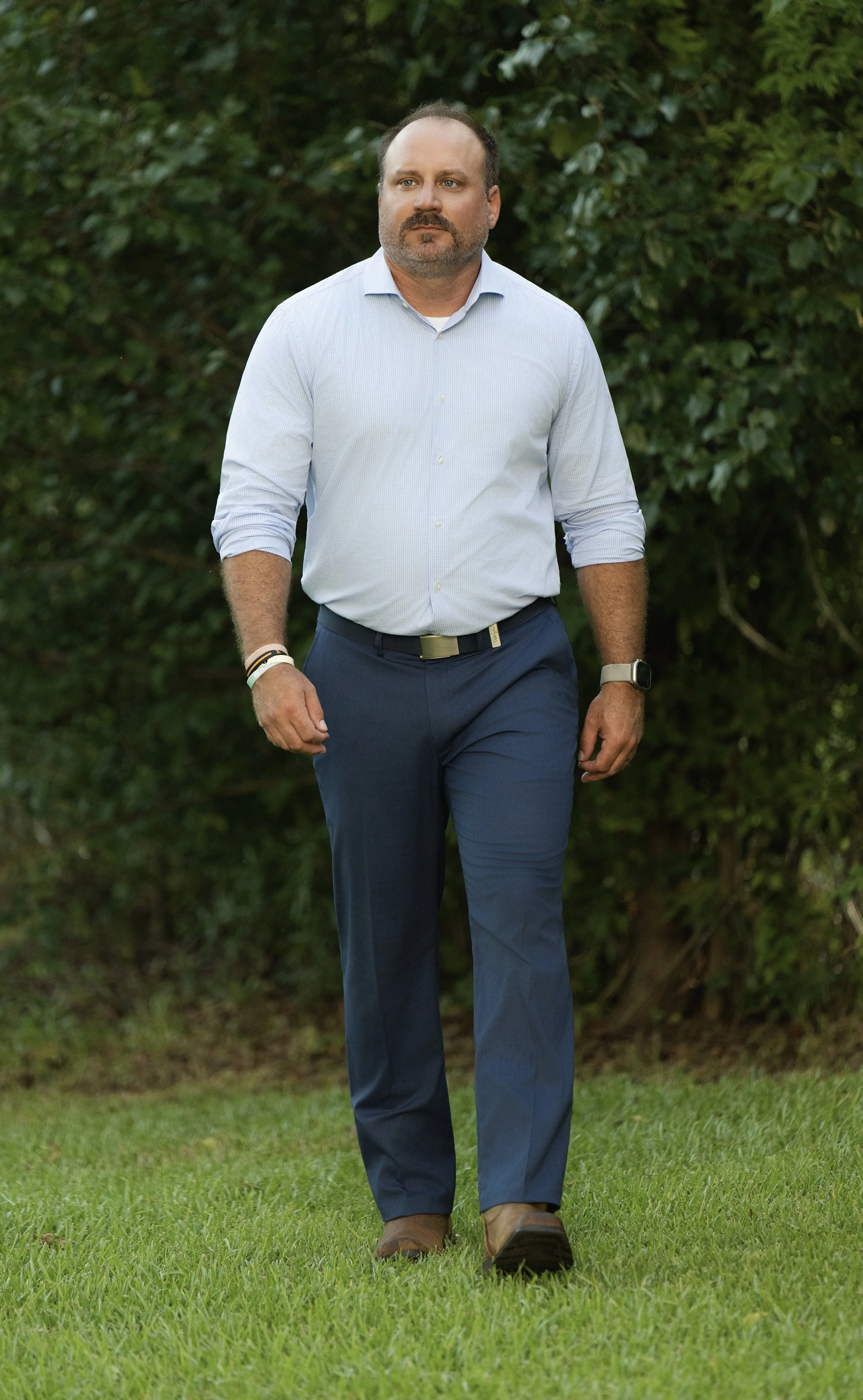 A man walking outdoors on grass with trees in the background, wearing a light blue dress shirt, dark blue pants, and brown shoes.