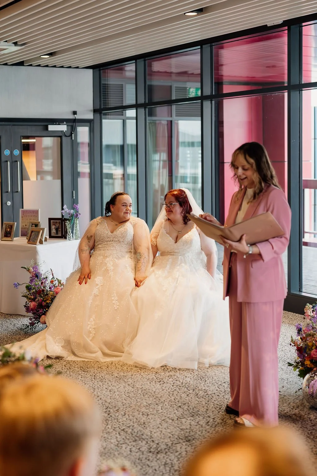 A lesbian couple, both wearing white dresses, smile at each other and hold hands as Hannah conducts their wedding