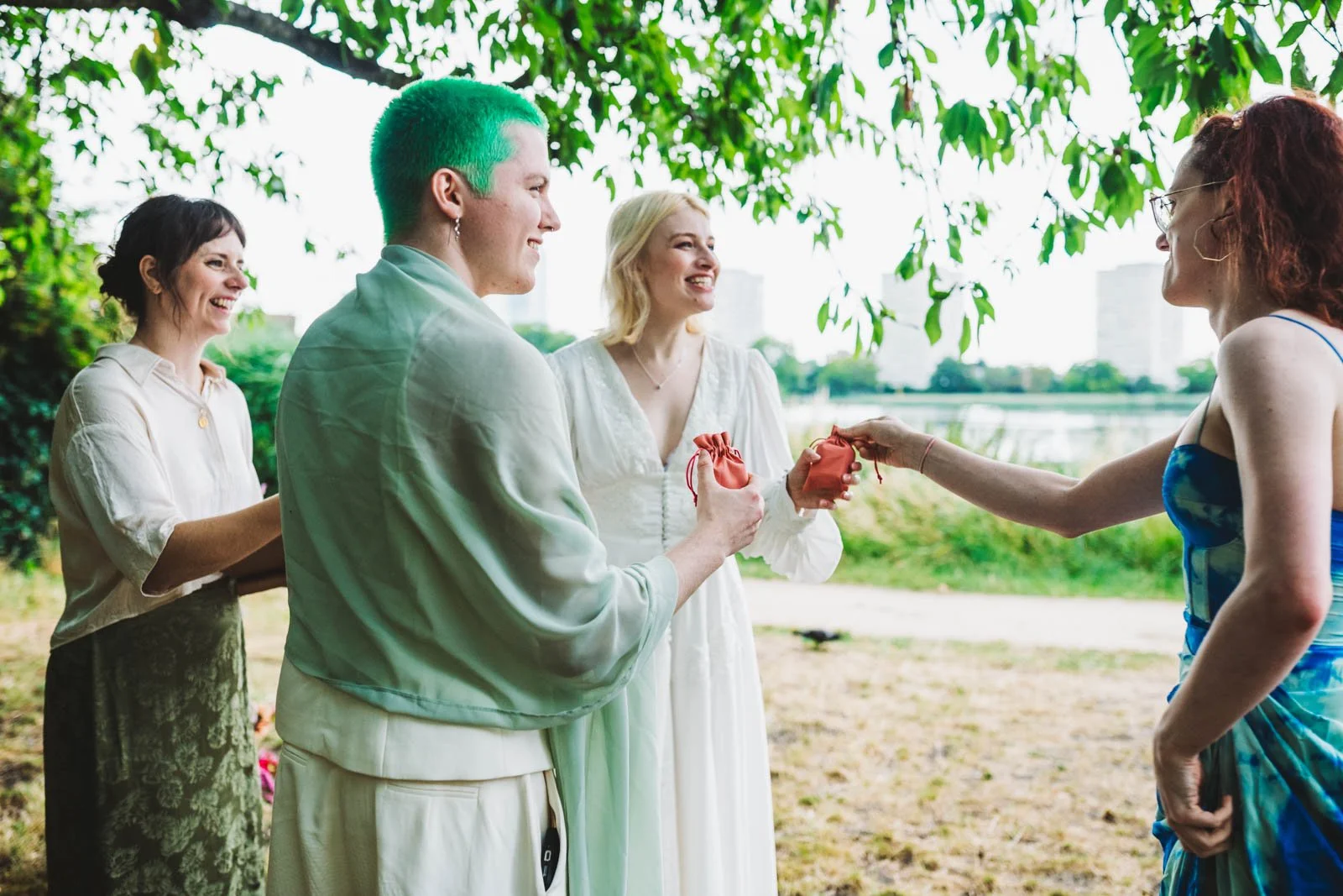 Hannah is standing outside with a non-binary person with green hair and a blonde woman in a white dress. They are being handed their wedding rings by a friend, everyone is smiling