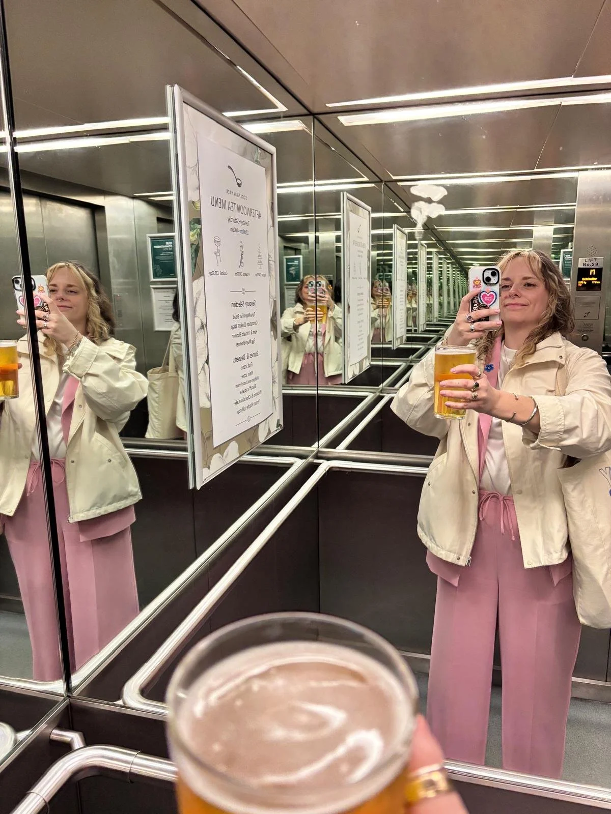 Hannah is taking a selfie in a lift mirror, smiling and holding a pint of lager up to the camera