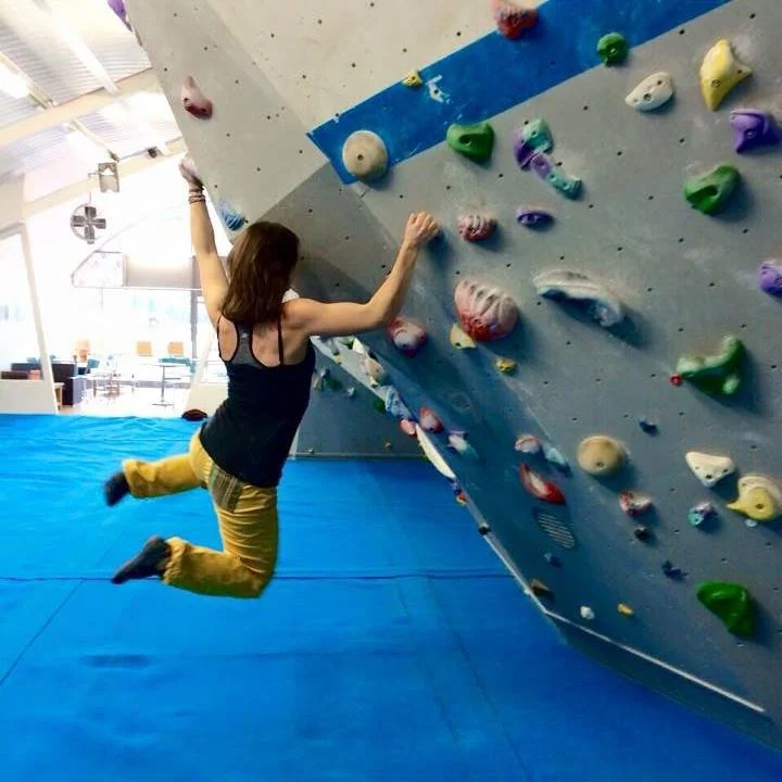 Hannah is hanging off of an indoor bouldering wall, legs swinging in the air