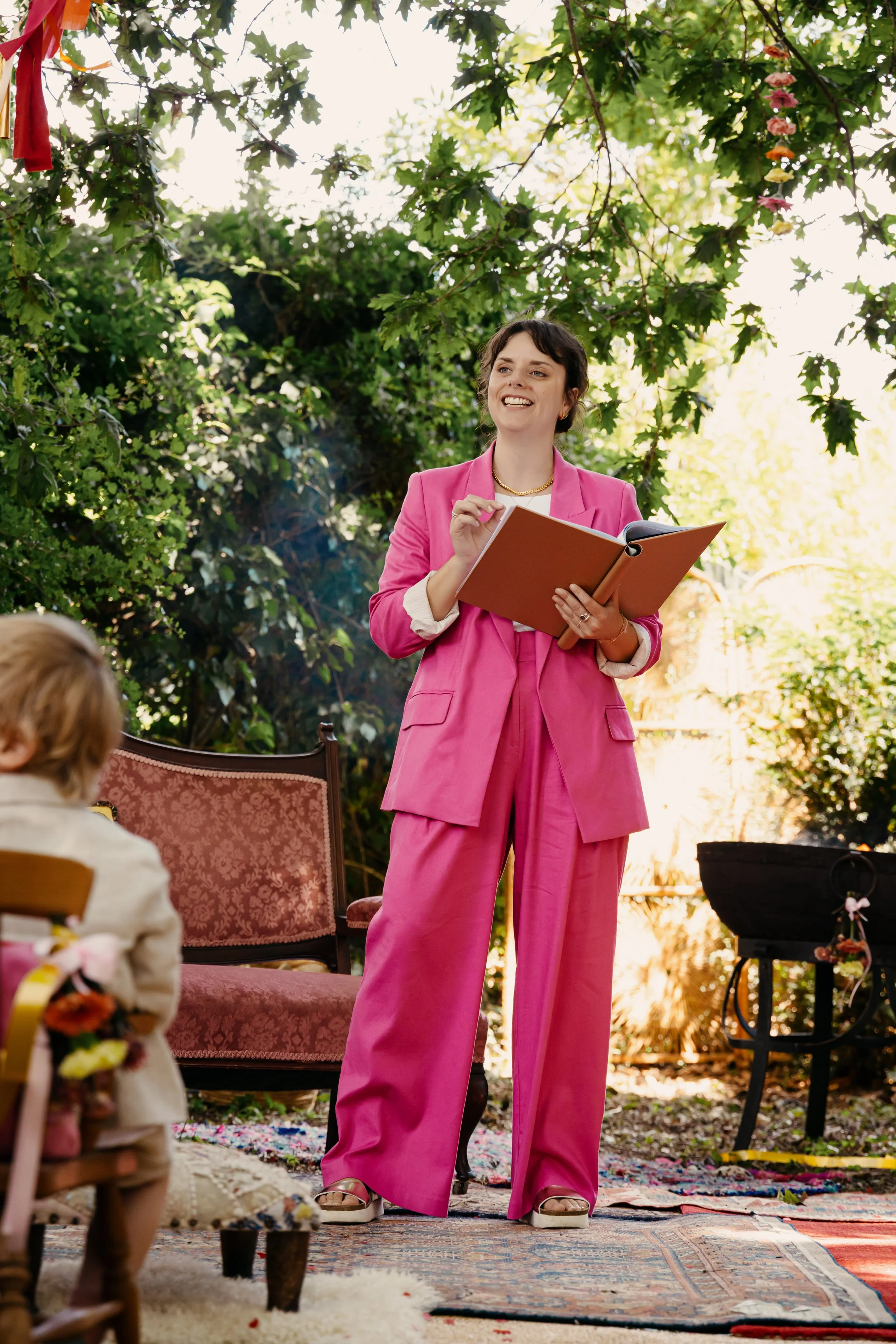 Hannah, a white woman in her 30s with brown hair and wearing a pink suit, stands smiling whilst reading from a folder during a ceremony