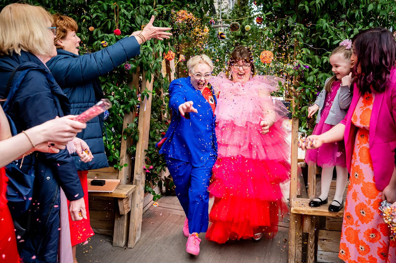 A non-binary/lesbian couple are laughing as they run under a cloud of confetti that has been thrown. One is wearing a bright blue suit and pink trainers, and the other is wearing a poofy red and pink dress