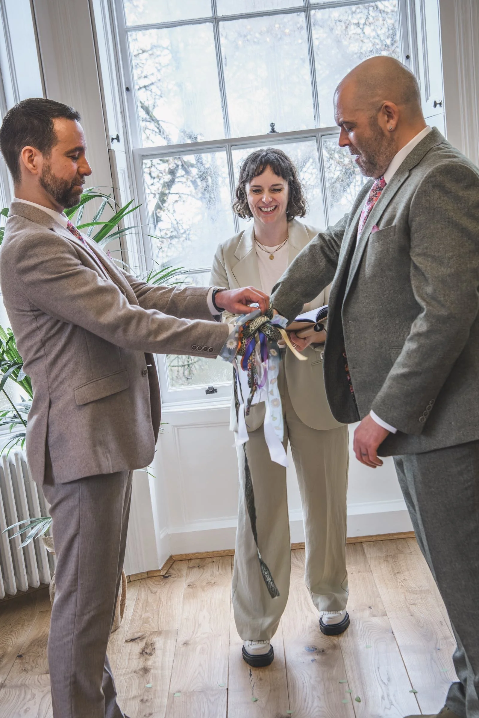 Hannah performs a handfasting for a gay couple, both wearing grey suits. Everyone is smiling