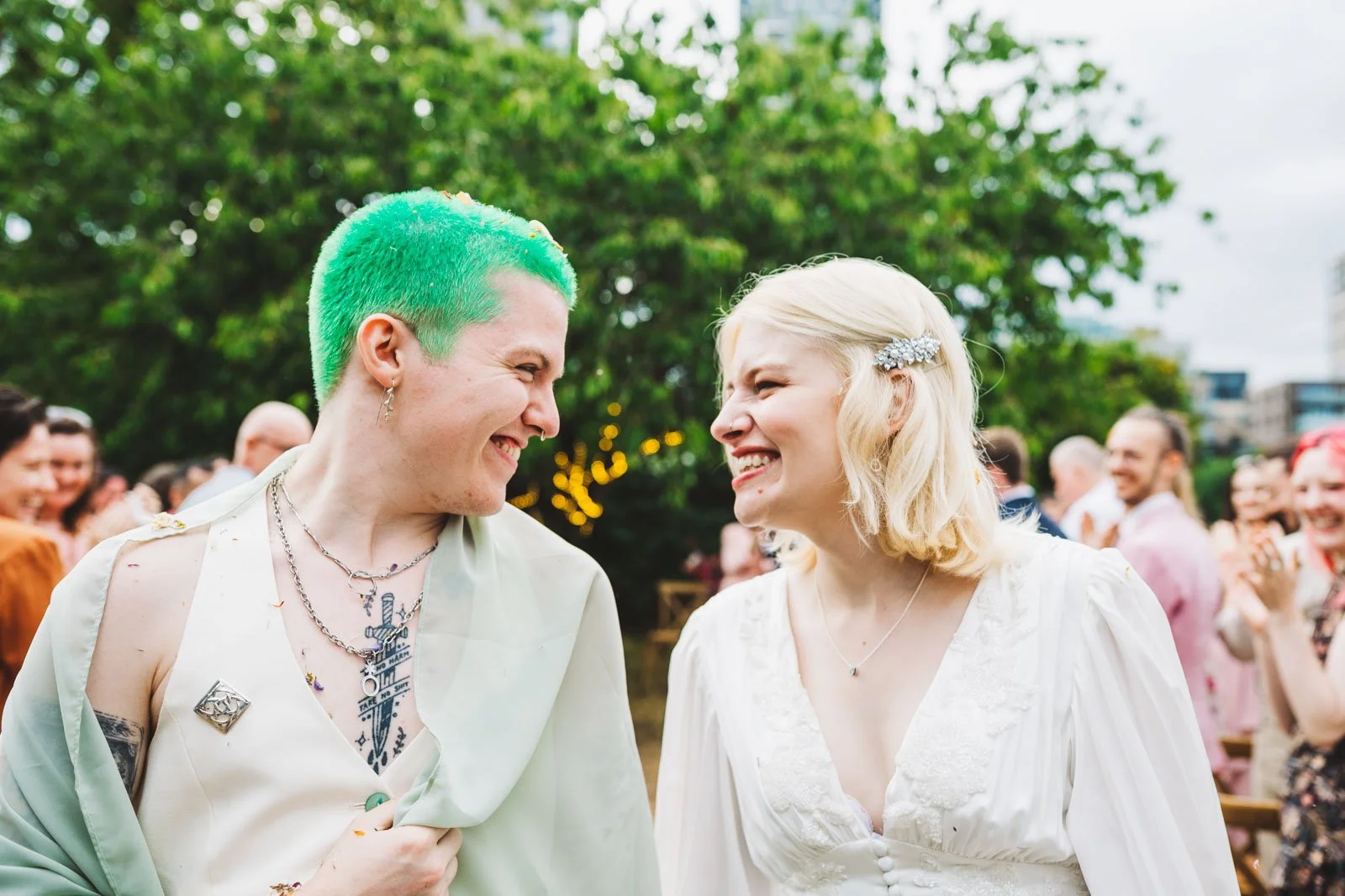 A non-binary person with green hair and a woman with blonde hair smile at each other broadly at their wedding