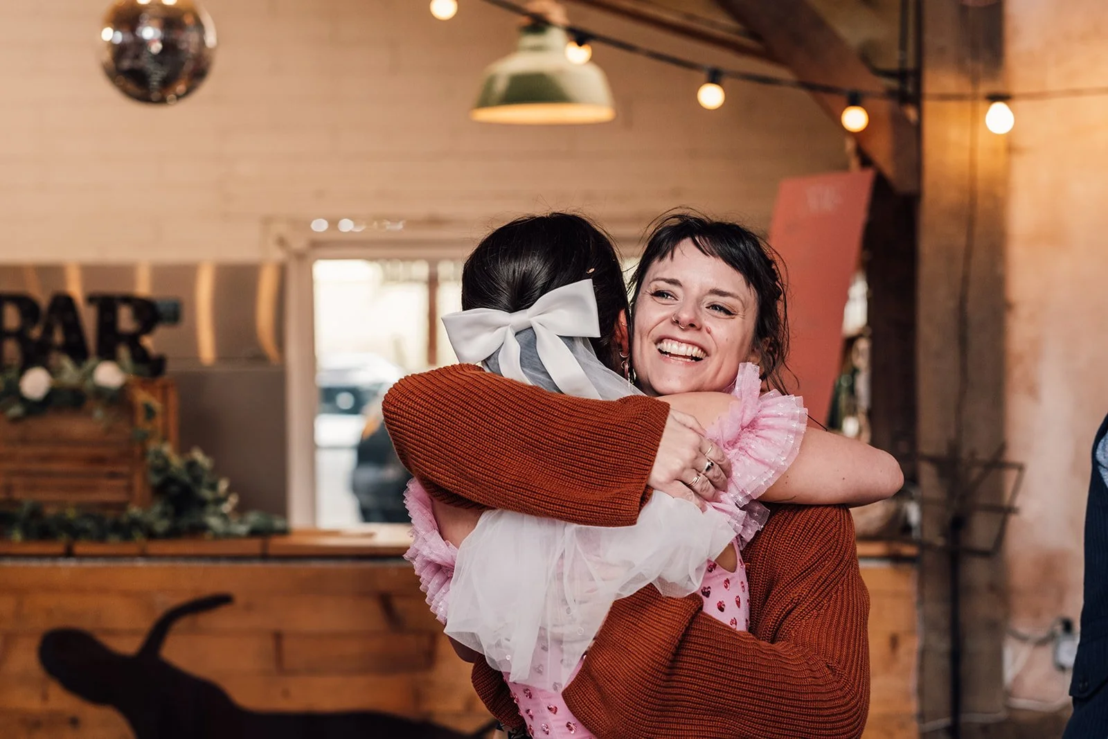 Hannah is smiling as she hugs a woman with brown hair, a large white bow, and a pink wedding dress