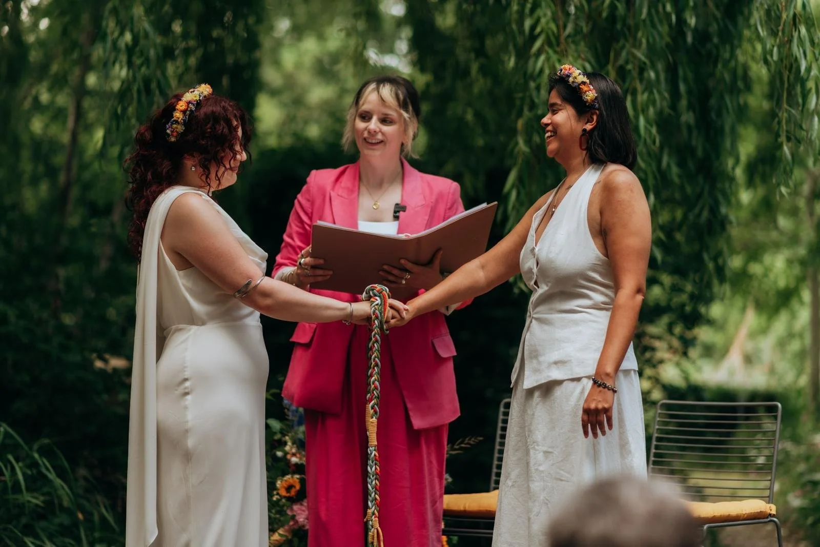 A lesbian couple hold hands as Hannah performs a handfasting. Everyone is smiling widley