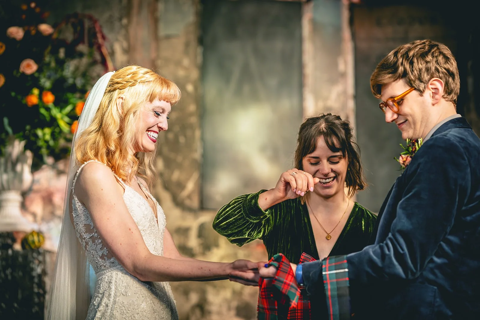 Hannah is performing a handfasting for a blonde woman in a white dress and a brunette man in a blue suit. They are all smiling broadly