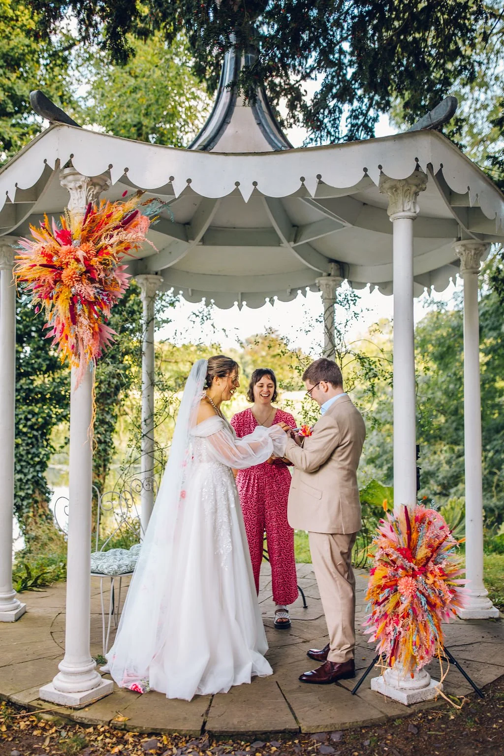 Hannah is standing under a white bandstand with a cis-het couple as they exchange their rings during their wedding, and everyone is laughing and smiling