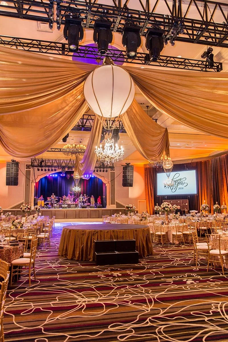 An elegantly decorated event hall with gold drapery, chandeliers, and a stage with live band performance, set for a celebration, possibly a New Year's Eve or anniversary event.