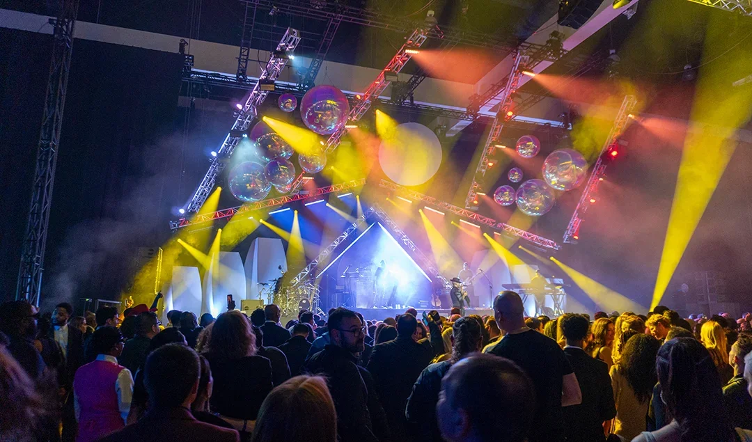 Crowd attending a concert with a stage, colorful lights, and large balloons overhead.