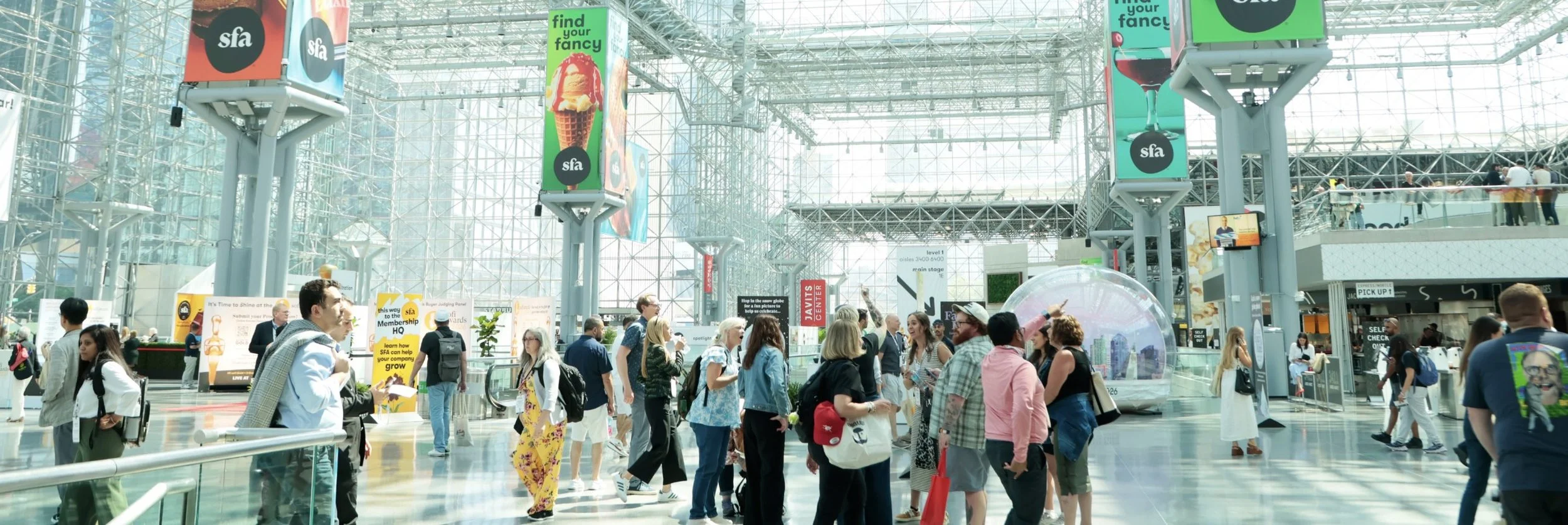 Crowd of people inside a modern glass-covered convention center or airport terminal with advertisements and signs hanging from the ceiling.