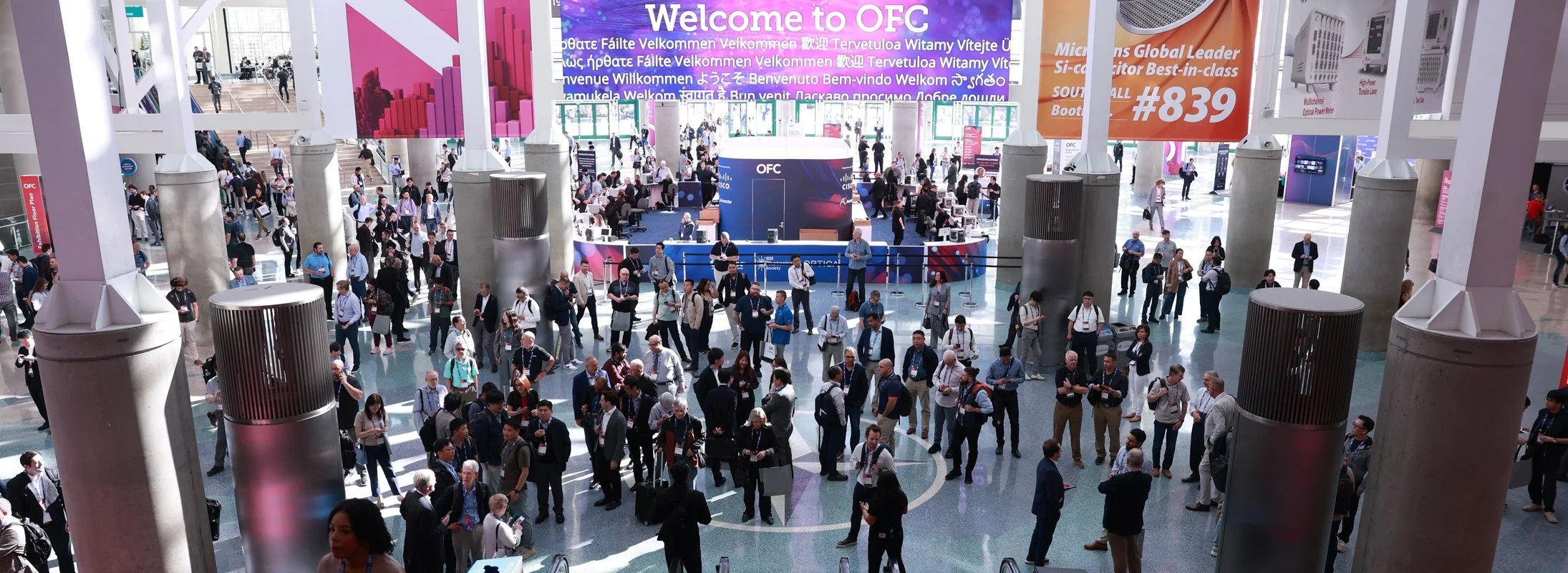 A crowded convention center with attendees walking and talking. There are large colorful signs overhead, including a prominent purple sign with white text that says "Welcome to OFC" and other signs relating to the event. The space features high ceili