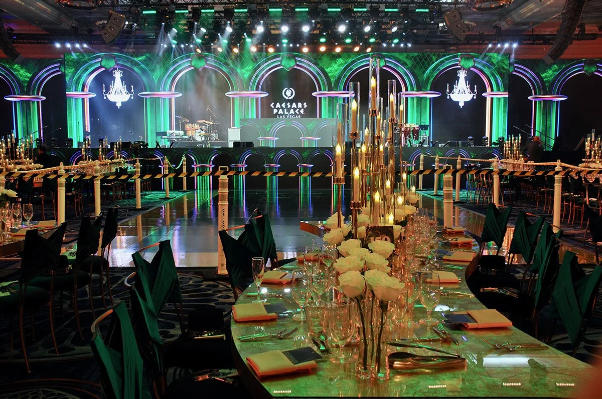 A large banquet hall decorated with elegant table settings, chandeliers, and stage lighting for an event at Caesars Palace in Las Vegas. The stage features colorful lights and electronic music equipment.