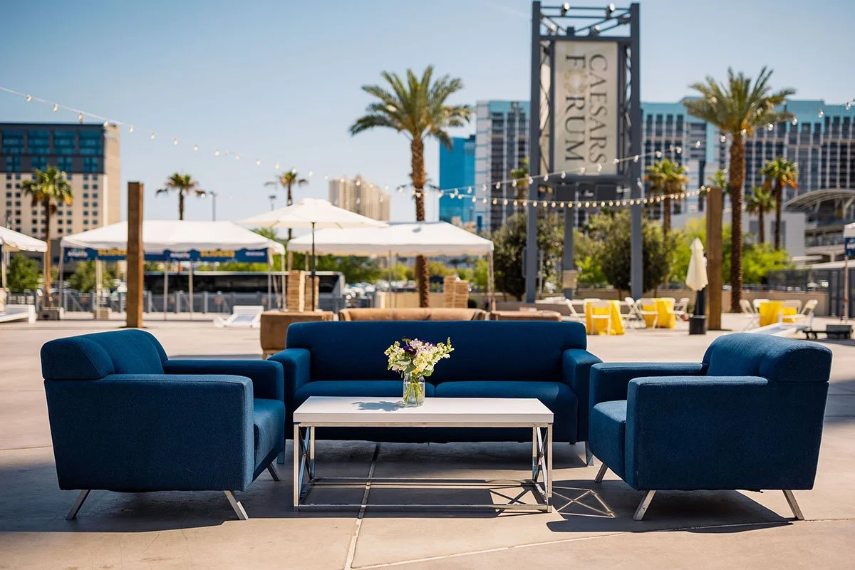 Outdoor seating area with three blue couches and a white table with a flower vase, Caesars Forum sign in the background, palm trees, and city buildings.