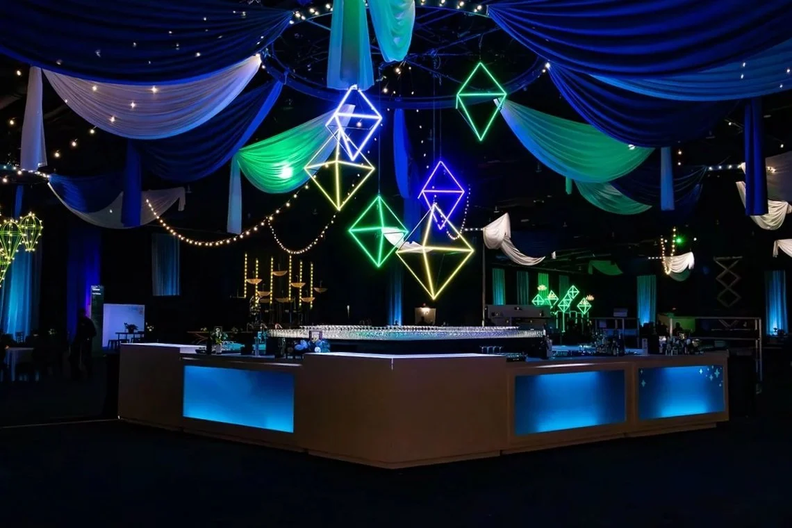 Colorful neon geometric light decorations hang from a dark ceiling decorated with draped fabric in blue, green, and white. String lights are also visible, creating a festive atmosphere in an event space or banquet hall.