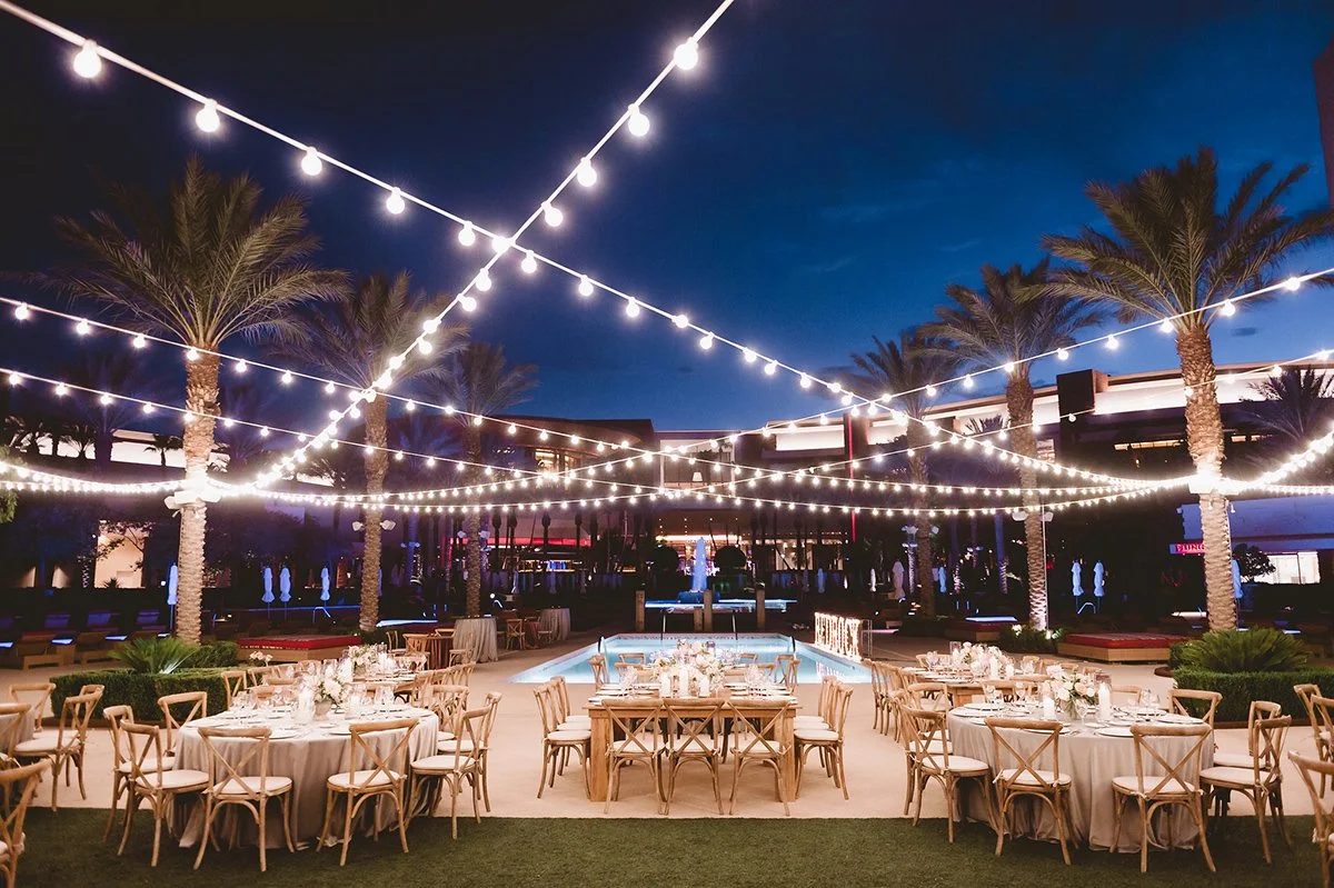 An outdoor event space at dusk with string lights hanging above, palm trees lining the area, and round tables with white tablecloths and floral centerpieces for dining.