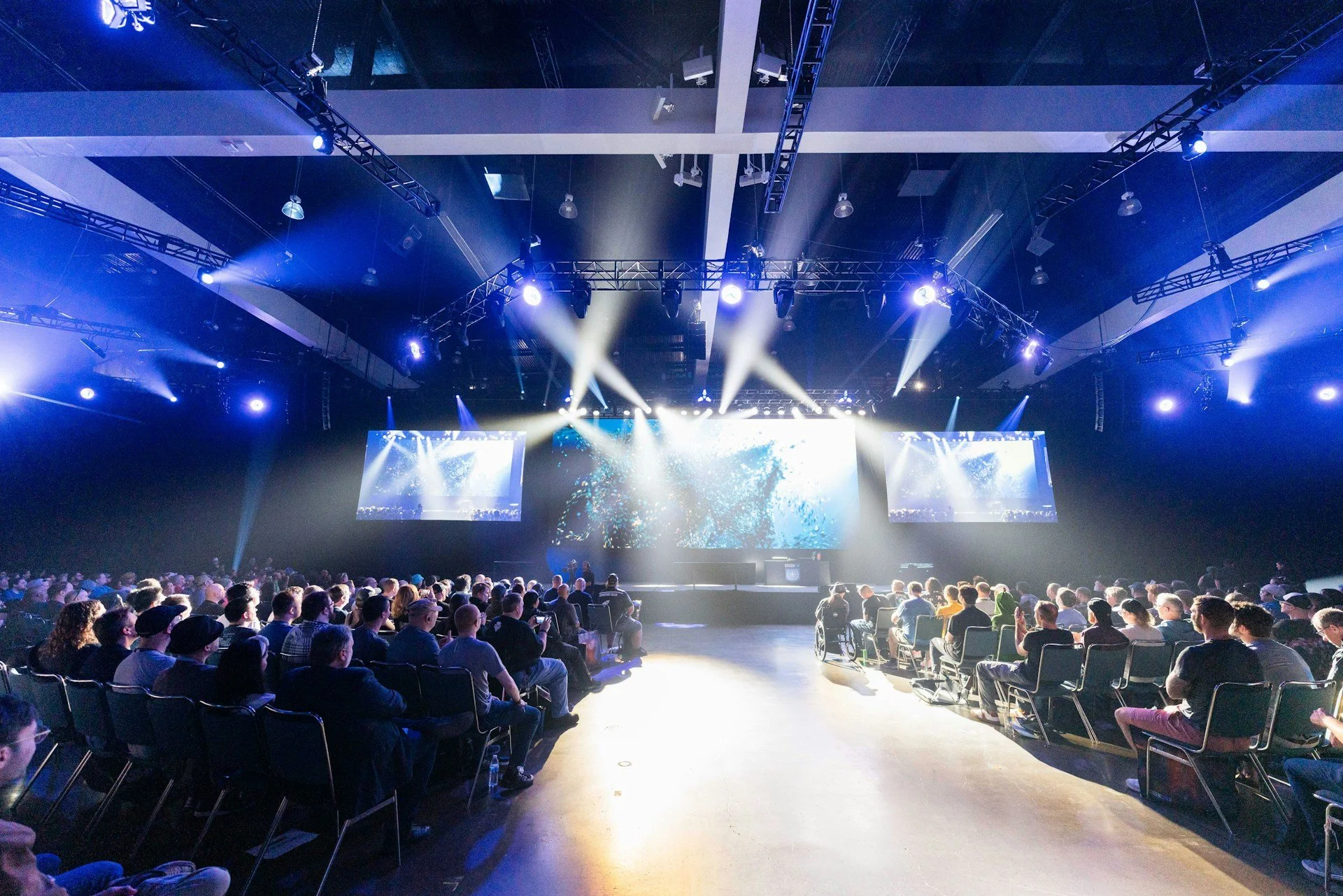 An audience seated in chairs facing a stage with large screens and bright lights in a dark indoor venue, likely for a concert, conference, or event.