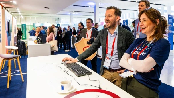 People smiling and engaging at a busy conference or trade show, with some using a computer at a white counter and others in the background exploring booths.